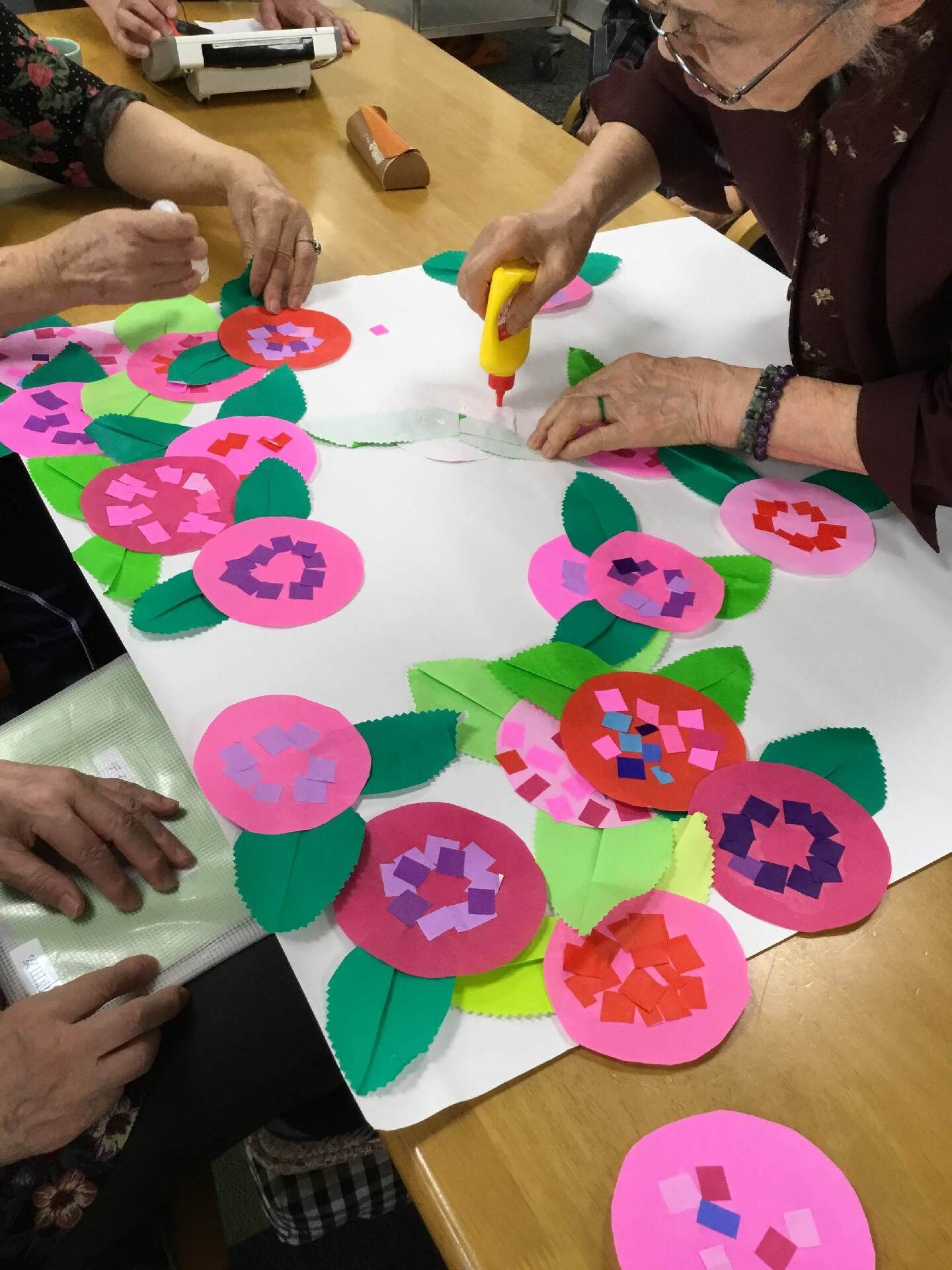 ピンクや赤の花を白い大きな画用紙に貼っていっている女性入居者たち。ボンドを出している女性入居者は眼鏡を少しずらしている。
