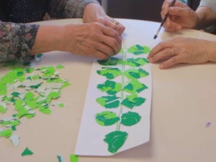 二人の女性入居者が長い葉っぱ下記のようなものをちぎり絵でしている様子。木緑と緑の折り紙を小さくちぎったものが紙の周りにある。