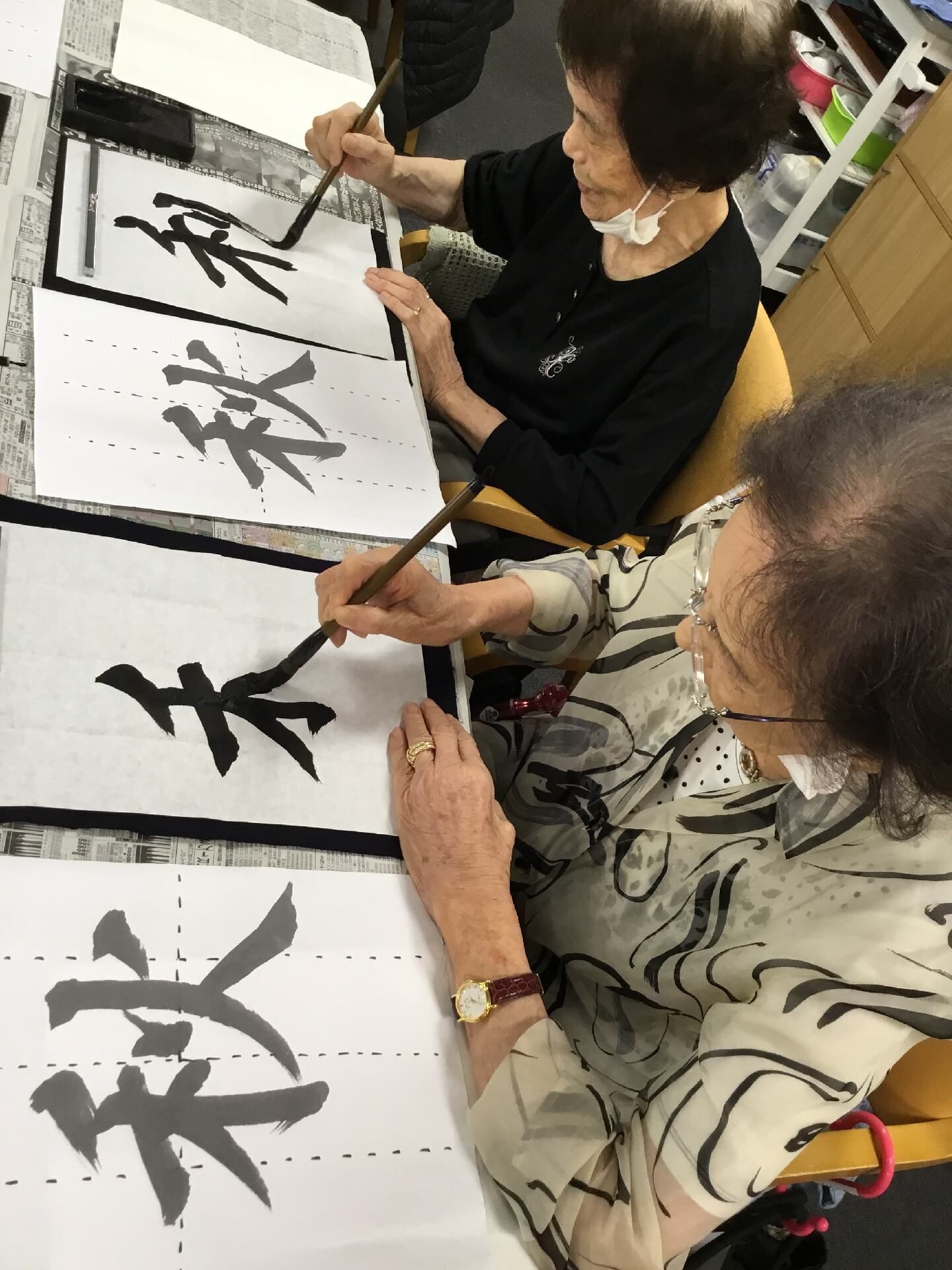 眼鏡をかけた高齢女性とマスクをした高齢女性がテーブルに並んで座り、書道をしている様子。「秋」という字を書いている。