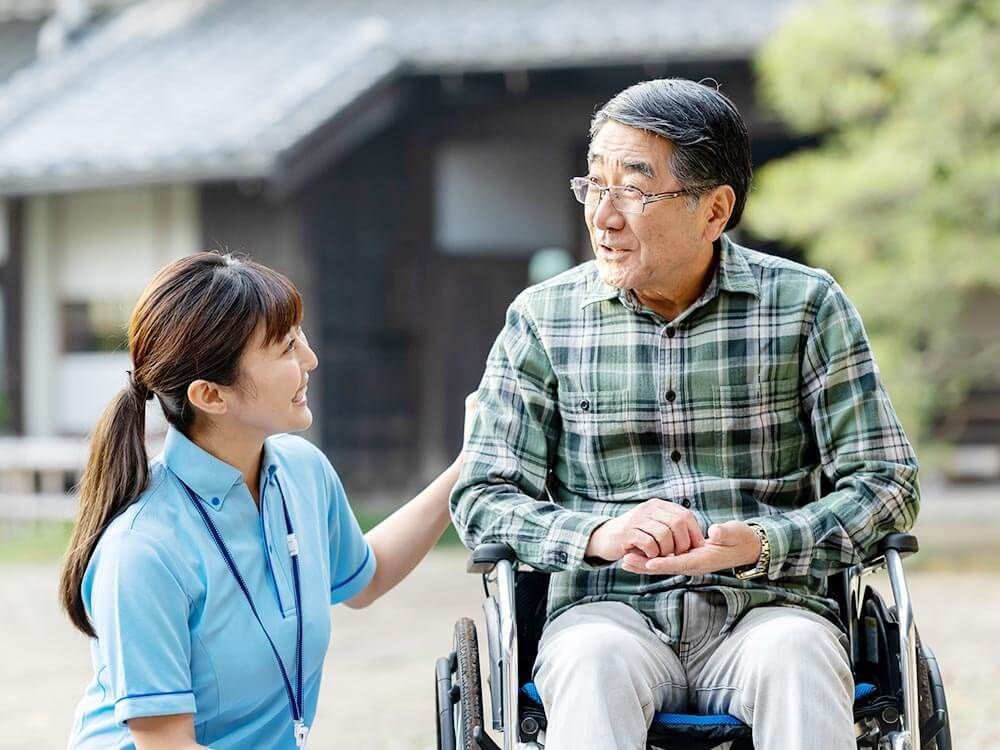 チェックのシャツを着た高齢男性が車いすに座り、そばにいるブルーのユニフォームを着た女性スタッフと話している。