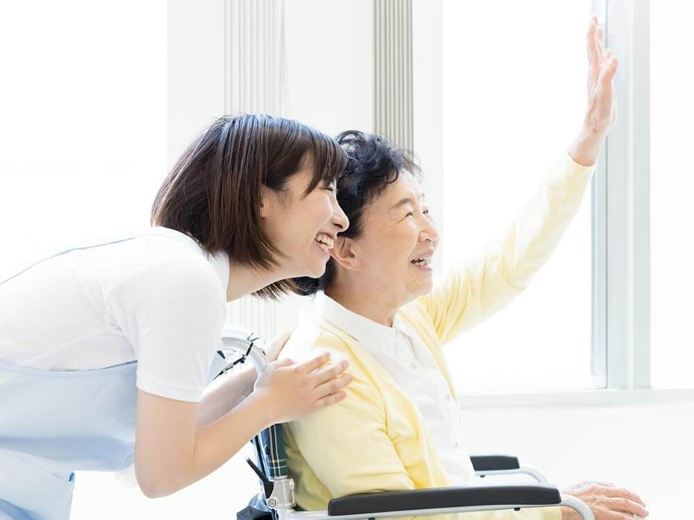 黄色いカーディガンを着た車いすの高齢女性が誰かに向かって笑顔で手を振っている。そばには女性スタッフがいる。