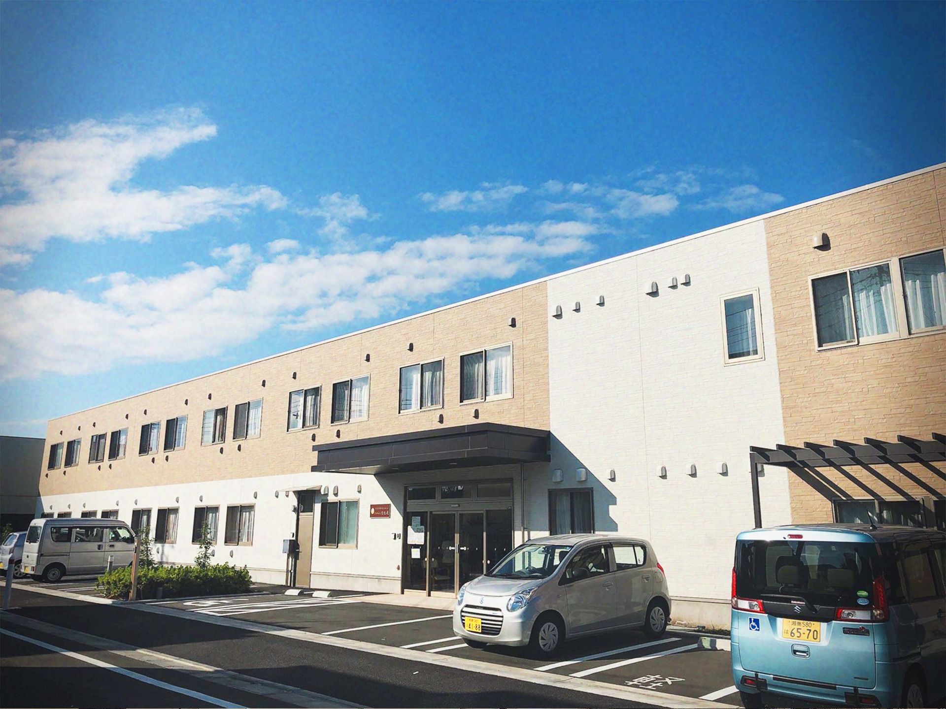 敷地内には駐車場があり、青い空の下に建っている2階建ての建物。外壁は白とベージュで、玄関に屋根がある。