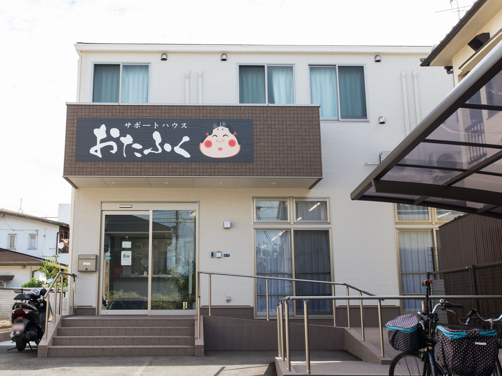 ２階建ての住宅が建っている。玄関前は手すりの付いた階段とスロープが設けられている。手前には屋根付きの駐輪場もある。