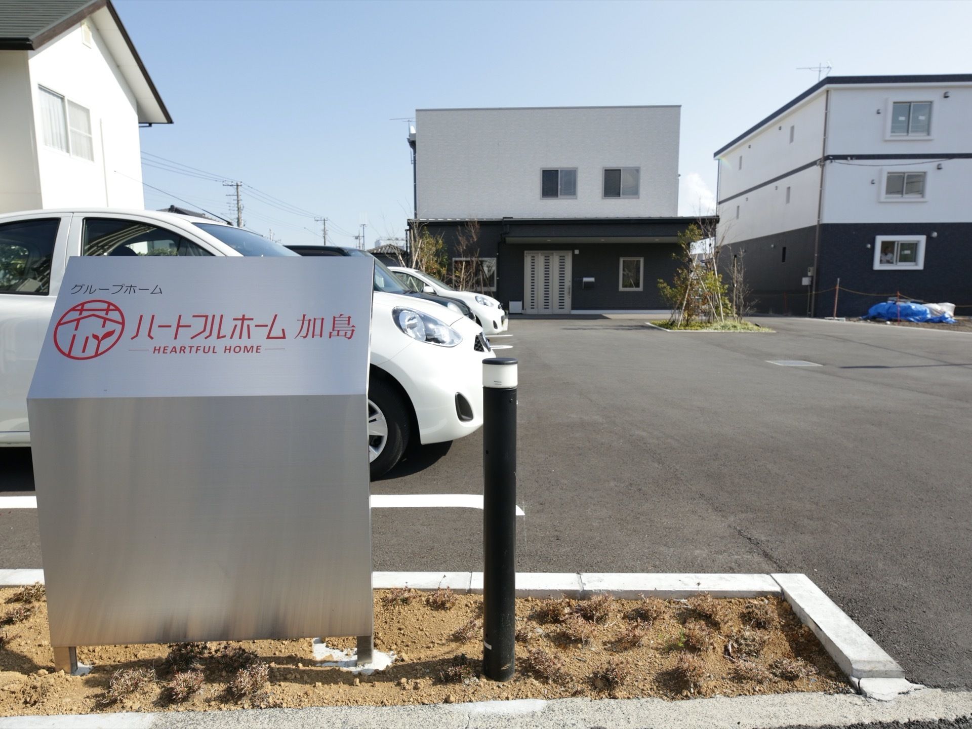 駐車場の入り口に施設の案内板が立てられている。金属板を曲げたデザインで、赤い文字でロゴと施設名が表記されている。