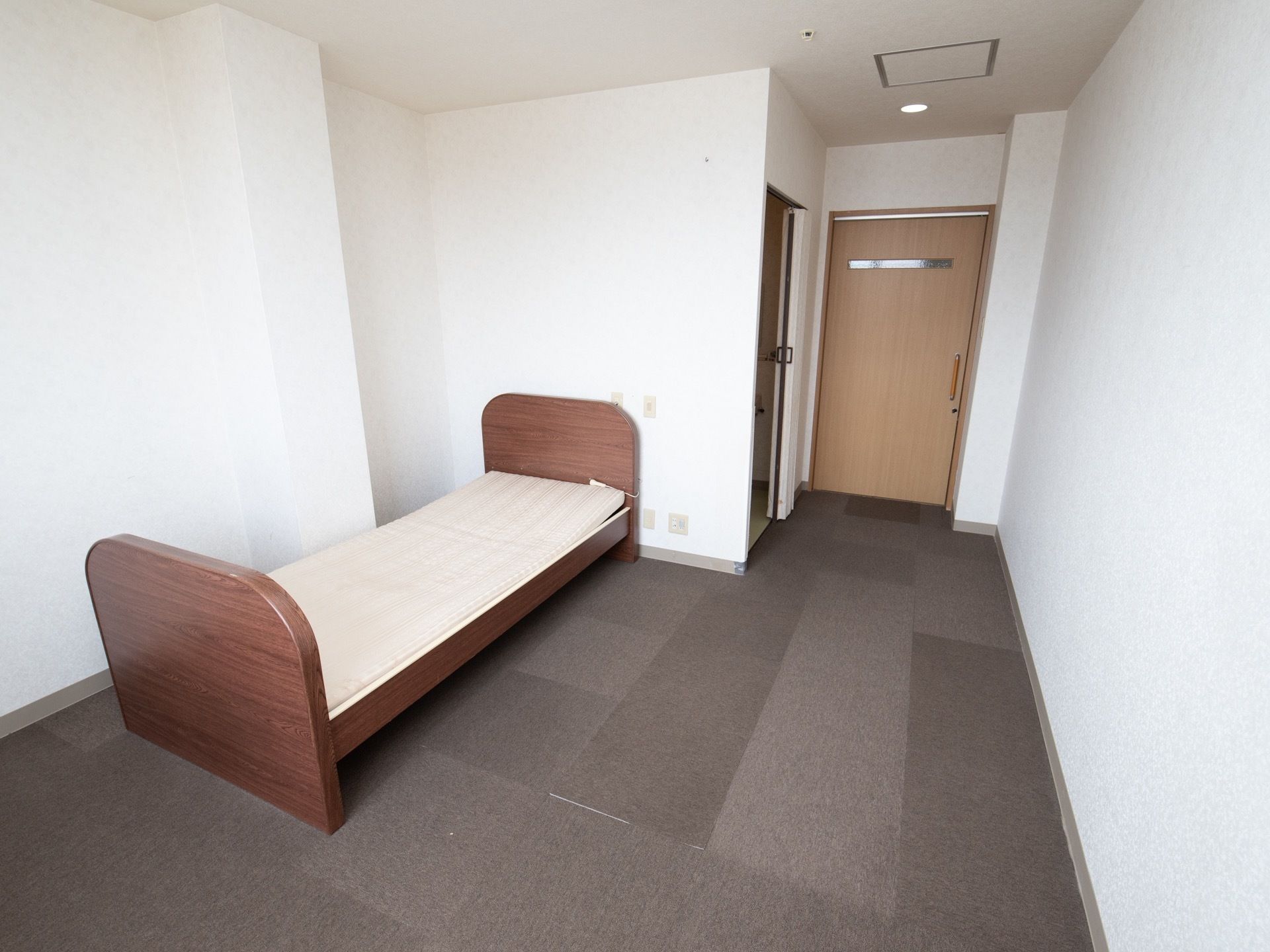 真っ白な壁が美しい広々とした居室である。介護用ベッドと玄関灯が完備されている。ダークグレーのカーペットが敷かれている。