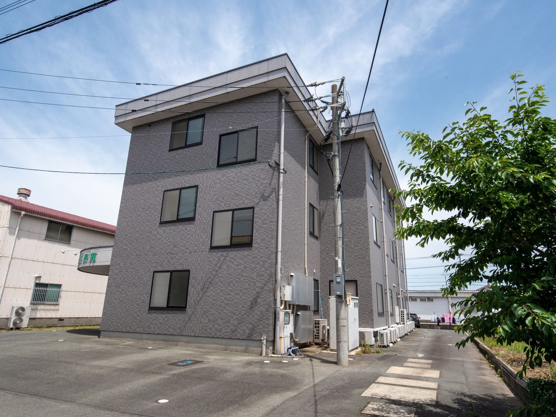 淡いシルバーのモダンな３階建ての建物である。周辺はアスファルトで整地された段差のない構造になっている。木々が茂っている。