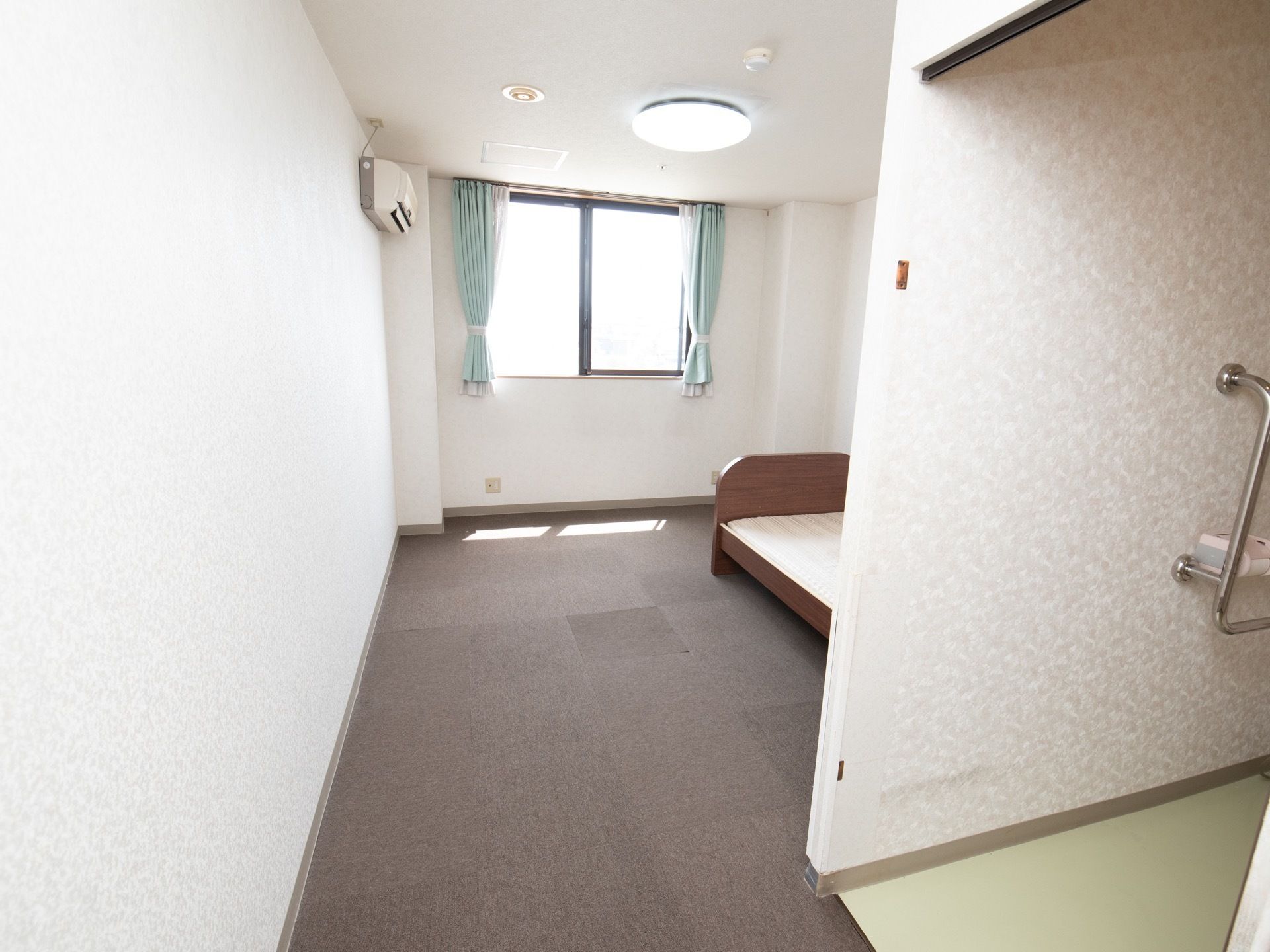 白が基調の明るい居室である。LEDライトやカーテン、介護用ベッド、エアコン、トイレが完備されている。