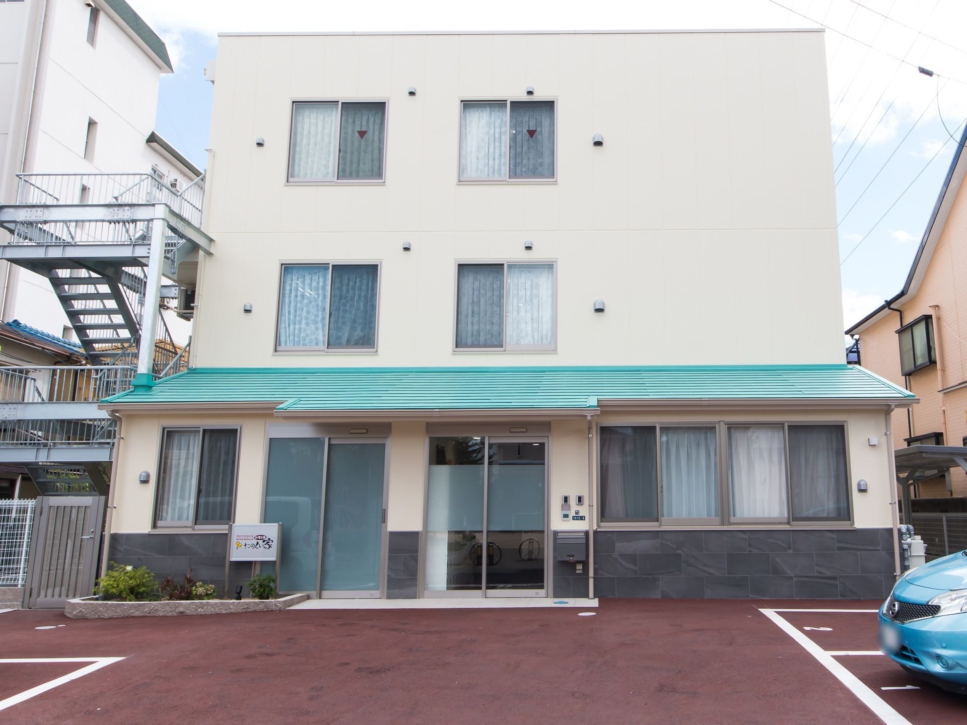建物を正面から見たところ。１階だけがやや前に出た構造になっている。敷地は赤く舗装されており、駐車場が設けてある。