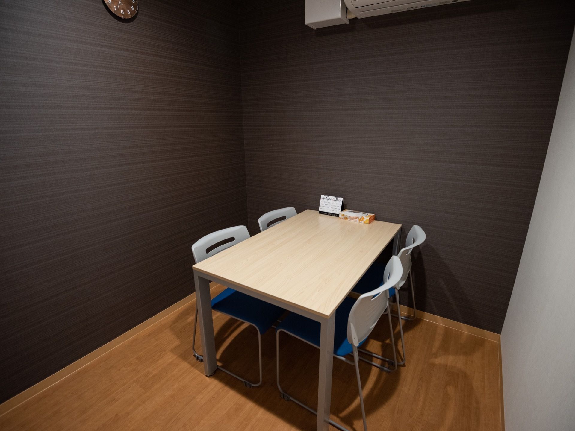 シックな色合いの壁の個室の部屋で、中央にテーブルと椅子を用意。テーブルの上にはカレンダーとティッシュ箱が置かれている。