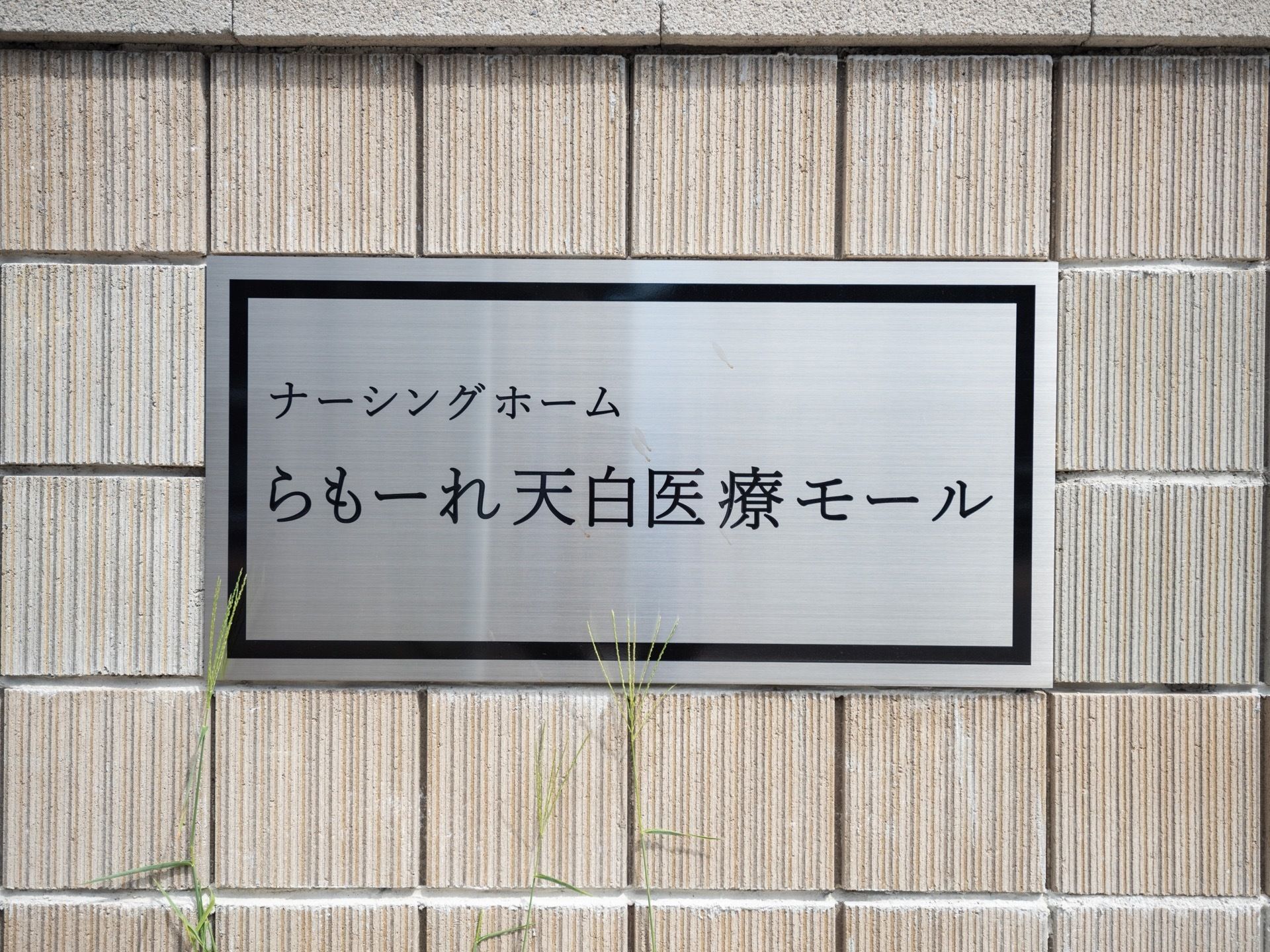 施設名が書かれた銀色のプレートが、外壁に埋め込まれる形で取り付けられている。プレートの高さまで植物が伸びている。