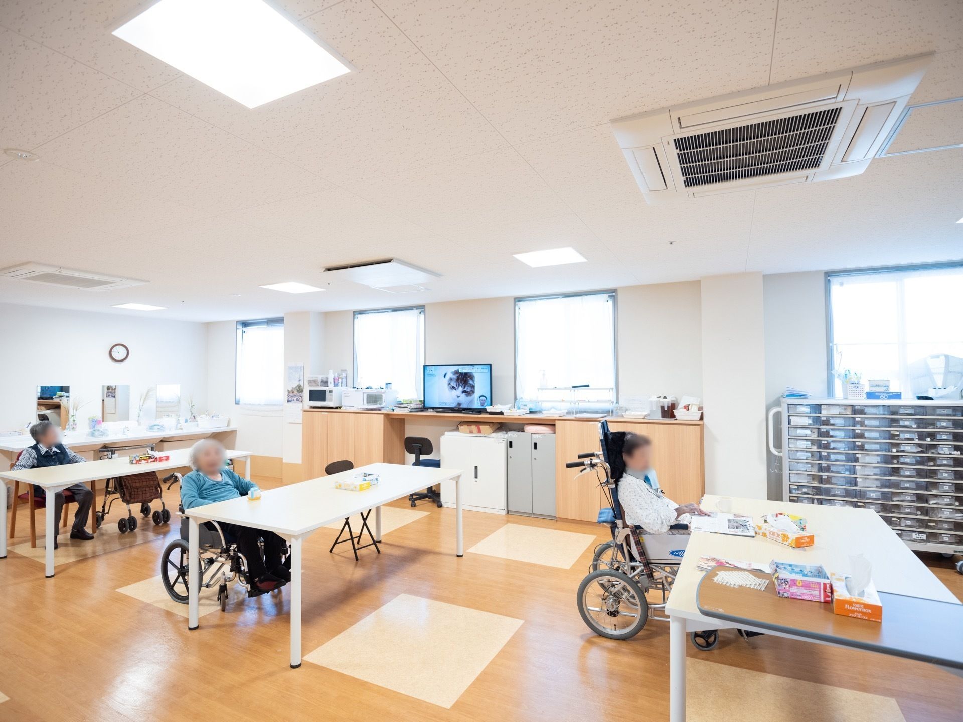 白い長テーブルが３台ある。入居者様が新聞を読んだり、テレビを観たりと、思い思いの時間を過ごされている。