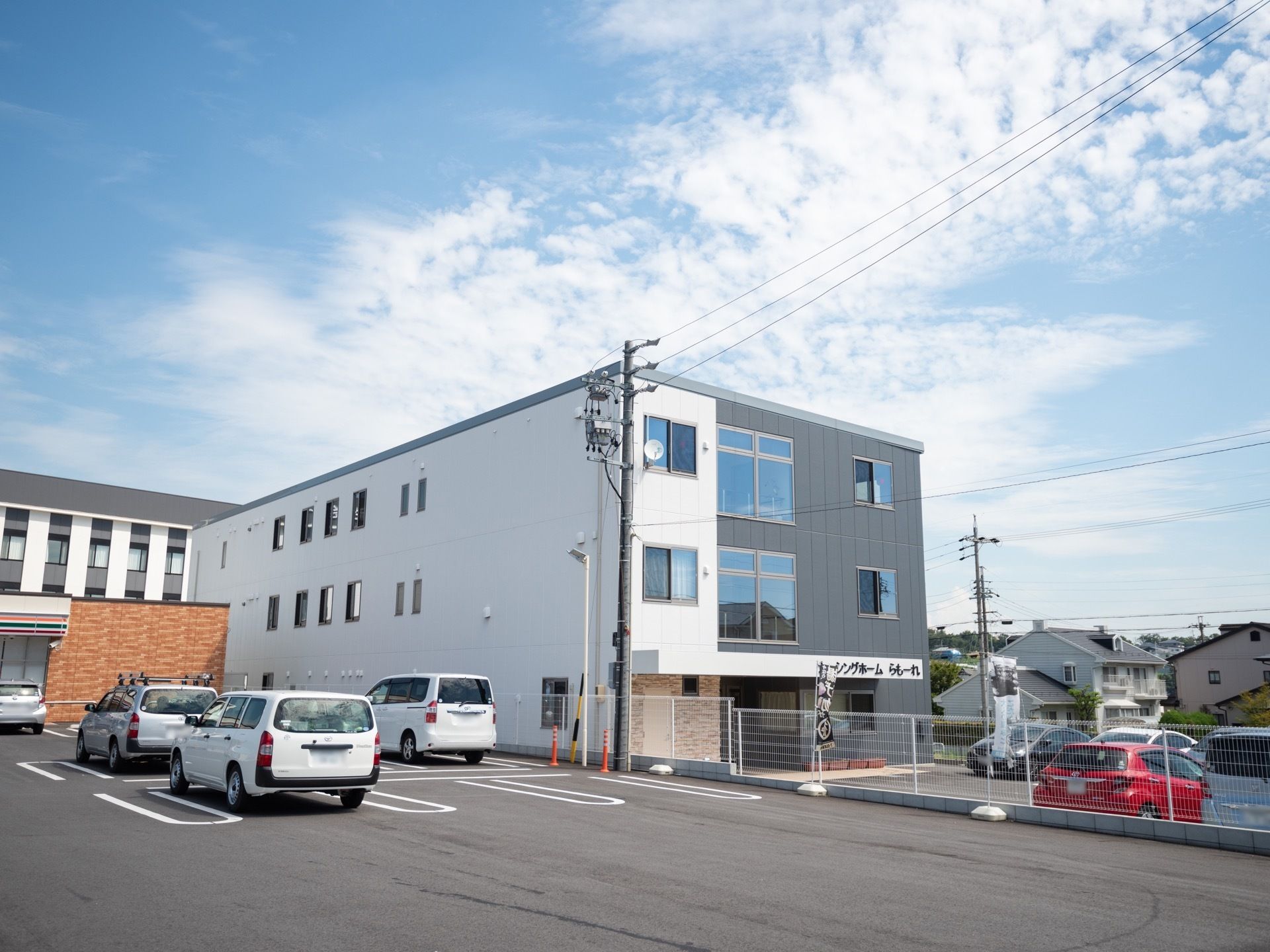 白とグレーの外壁の３階建ての建物で、施設前は駐車場になっている。隣にある商業施設とは白いフェンスで区切られている。