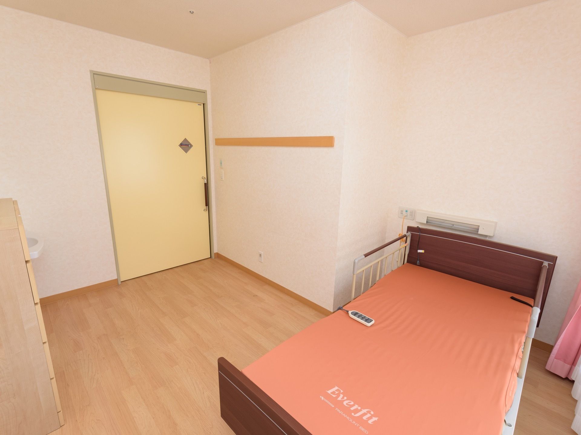 左手奥にクリーム色の扉の入口があり、引き戸になっている。手前には介護用ベッドが置かれ、リモコンが取りつけられている。