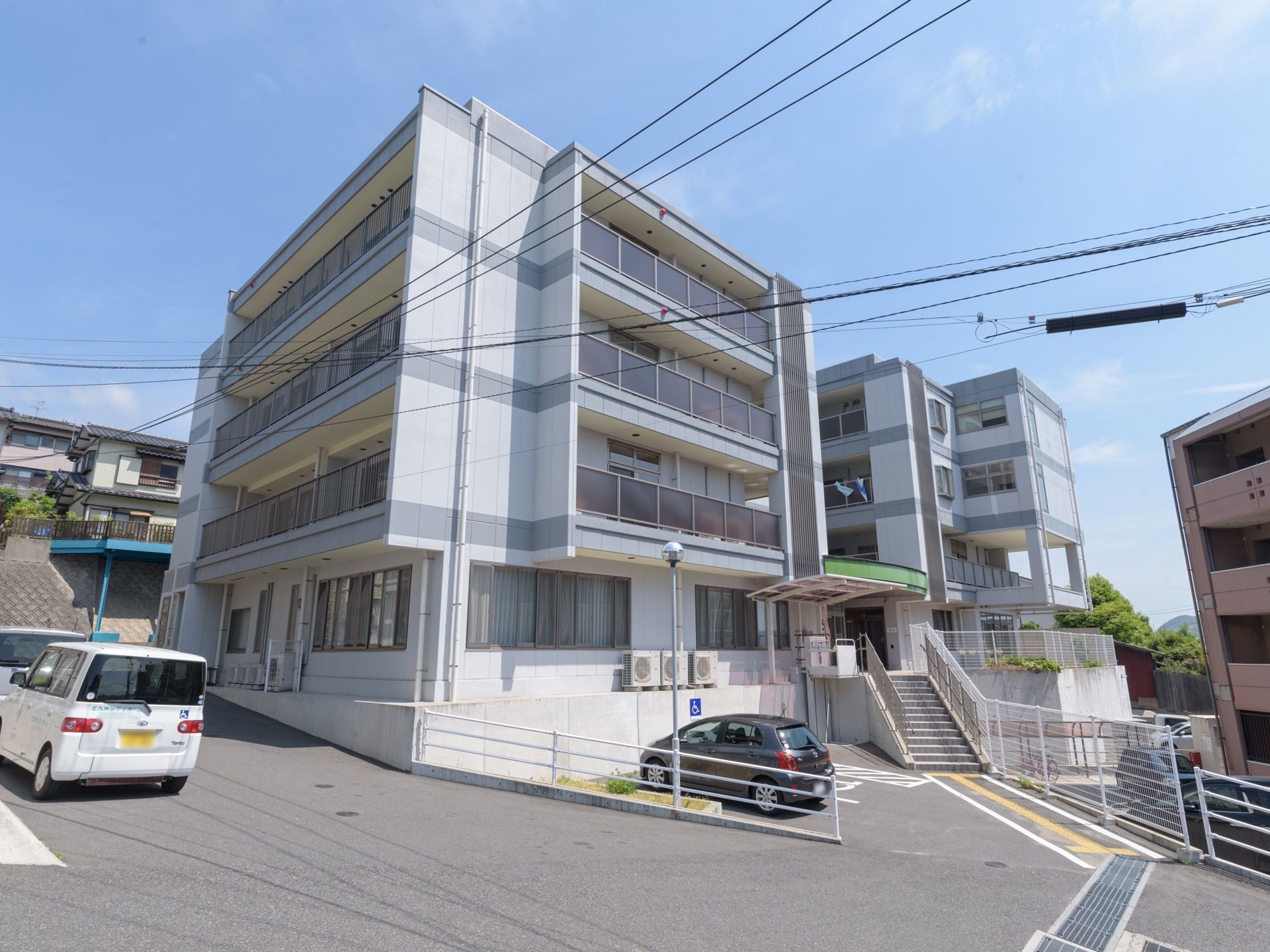 山の斜面に建つ４階建ての建物で、白を基調にグレーのラインが入っている。施設入口へは階段が伸びている。