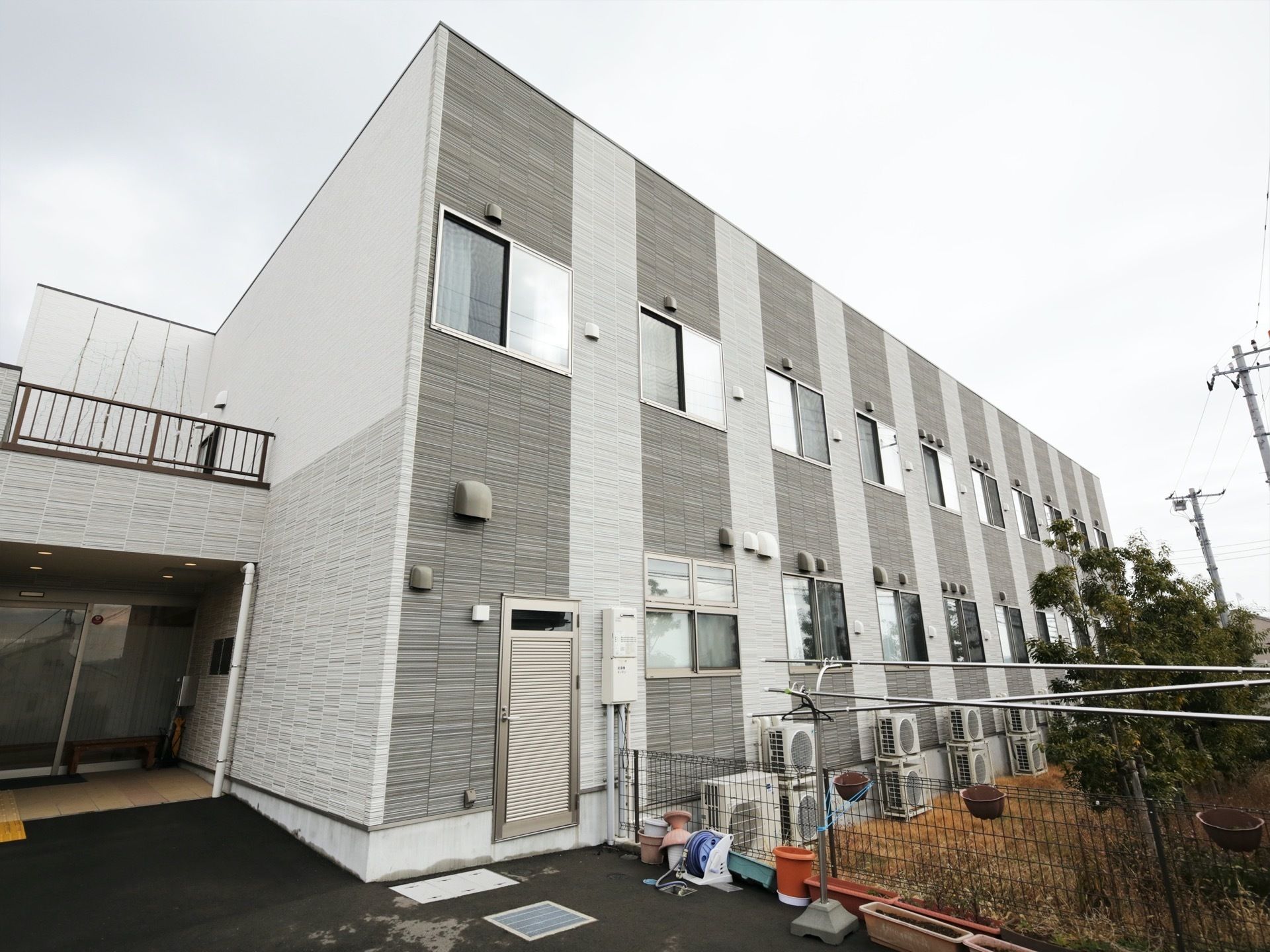 木造２階建ての施設で、グレーの外壁が落ち着いた印象である。各居室に設置されているエアコンの室外機が並んでいる。
