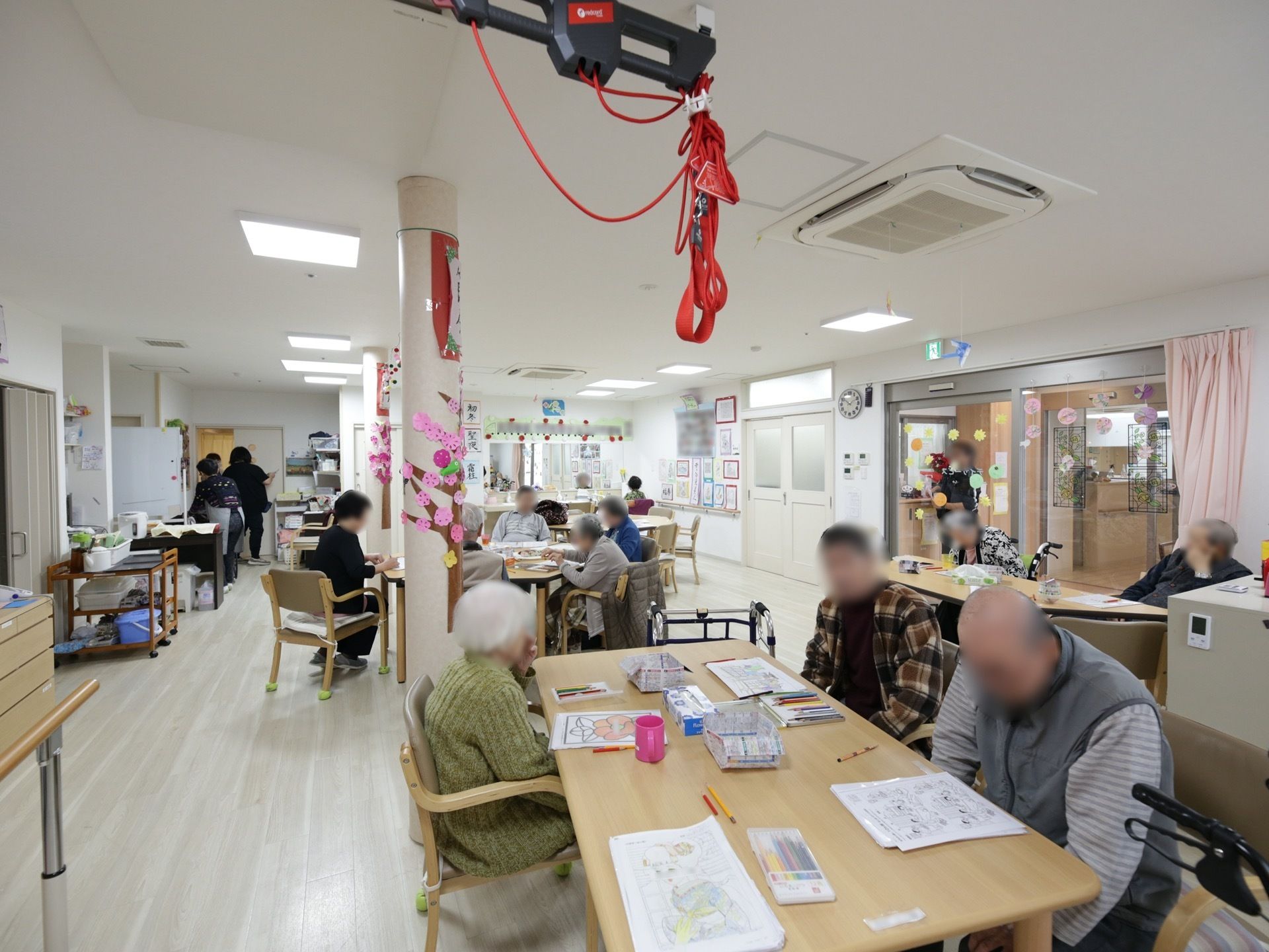 入居者様がテーブルにつき、それぞれ手作業をしている。奥にはスタッフの姿も見受けられる。部屋の隅に機能訓練機器が置かれている。
