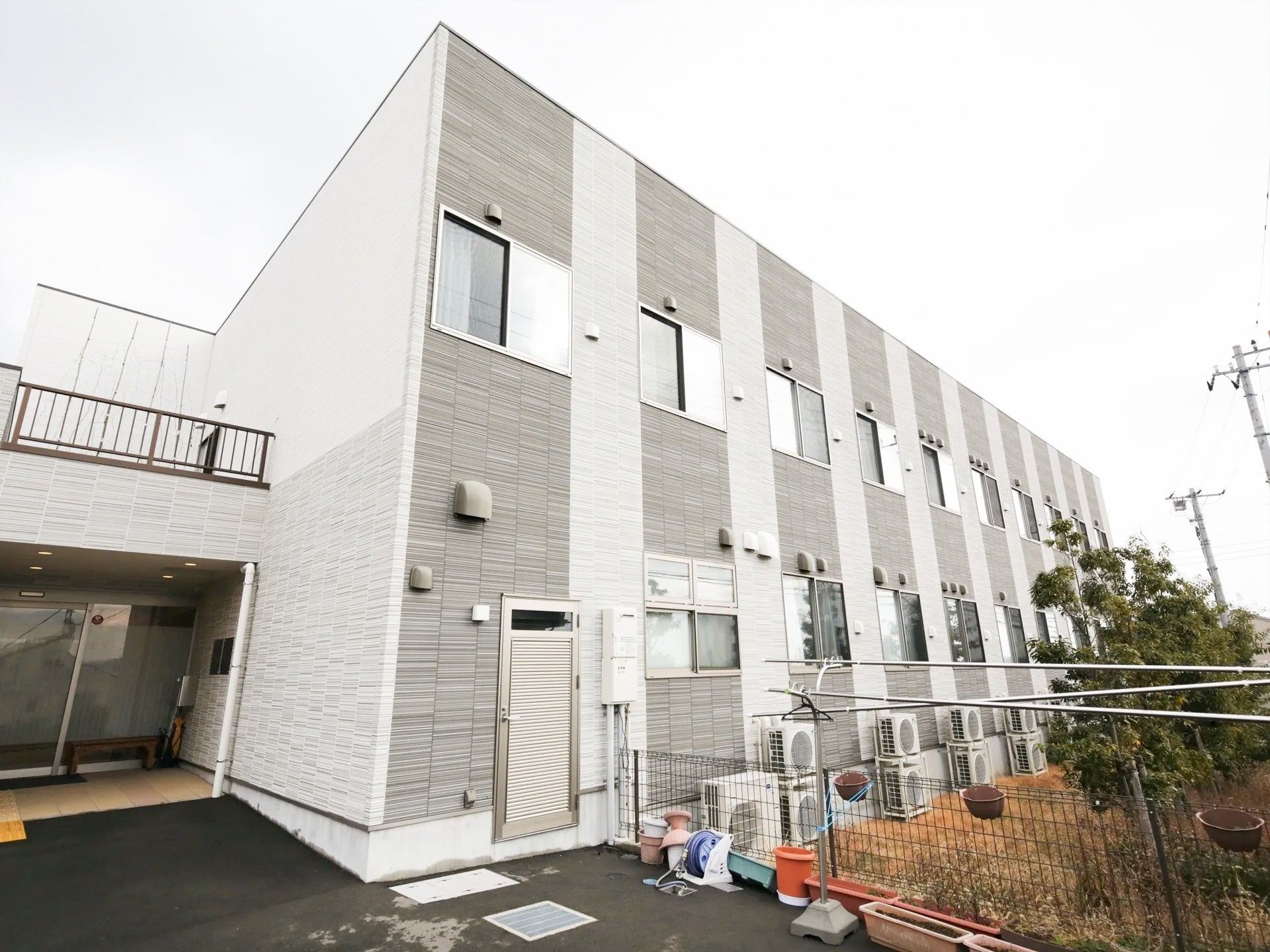 木造２階建ての施設で、グレーの外壁が落ち着いた印象である。各居室に設置されているエアコンの室外機が並んでいる。