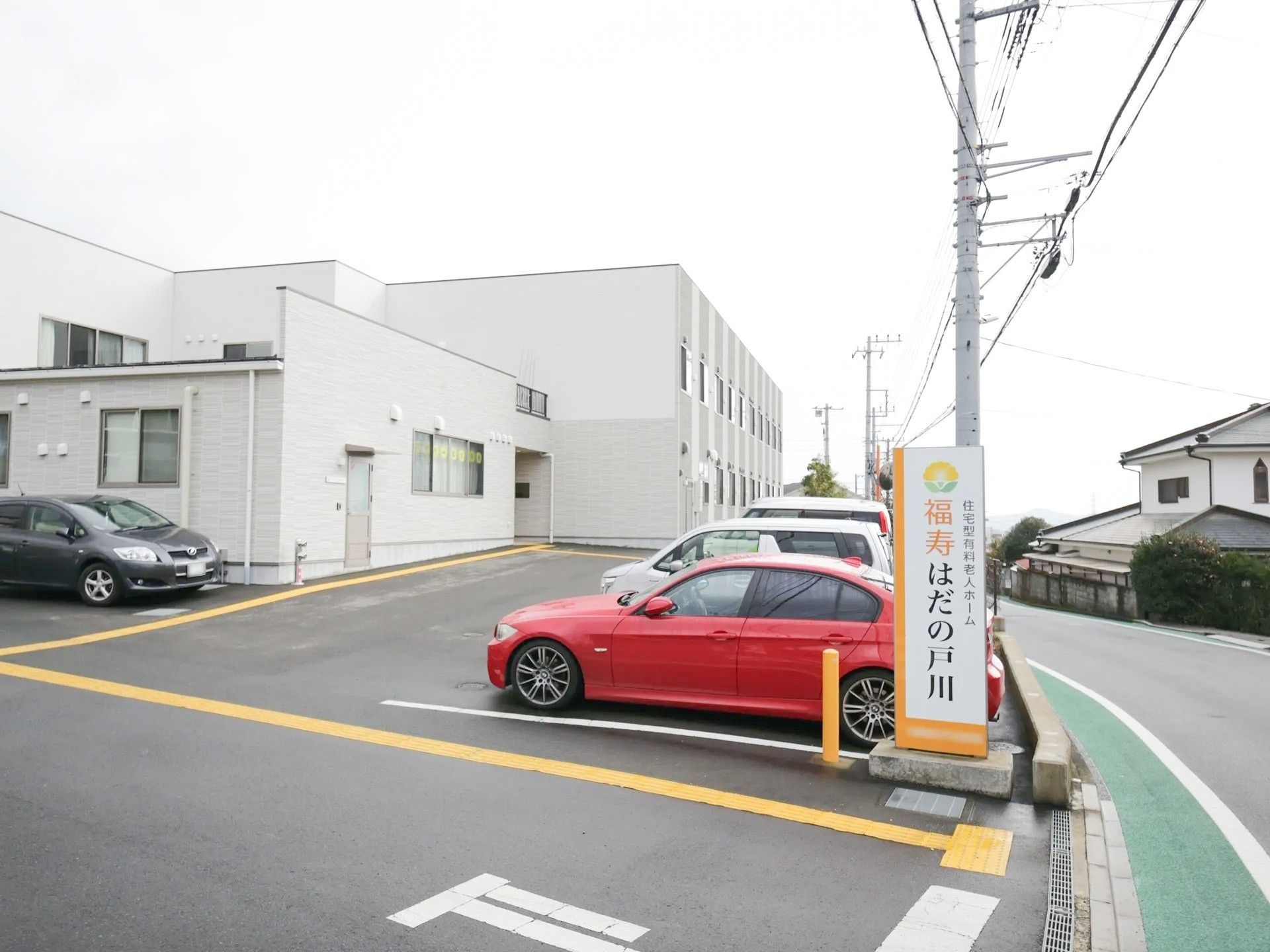 駐車場入り口に施設名の看板が設置されている。道路から見やすい場所にある。施設建物と駐車エリアは同じ敷地内にある。