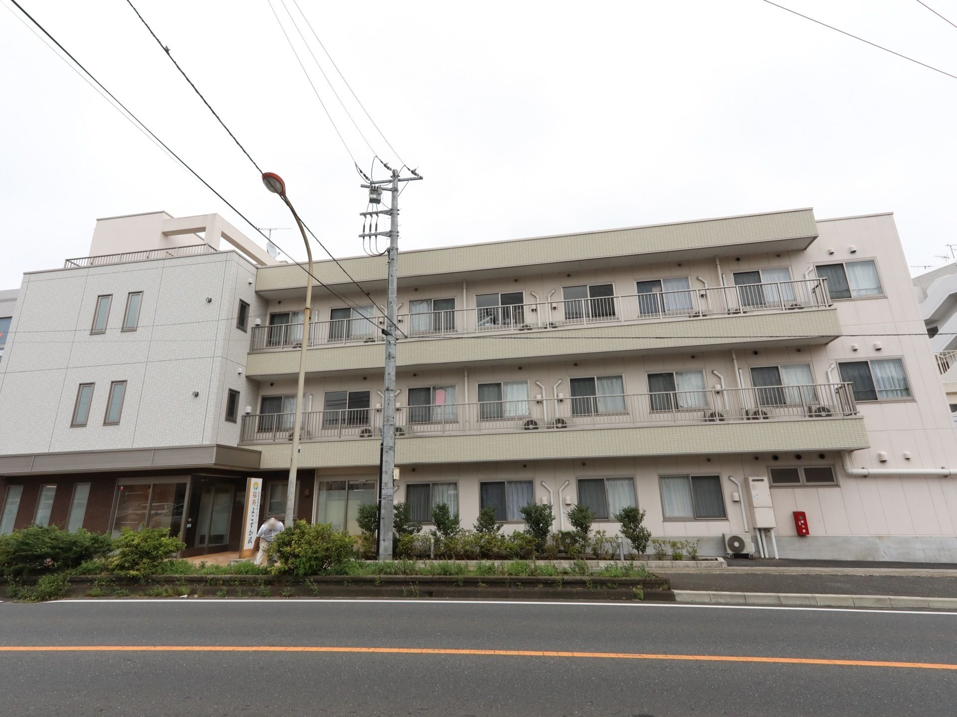 道路に面して建っている３階建ての施設である。玄関前に看板が立てられ、その横の歩道沿いには木が植えられている。