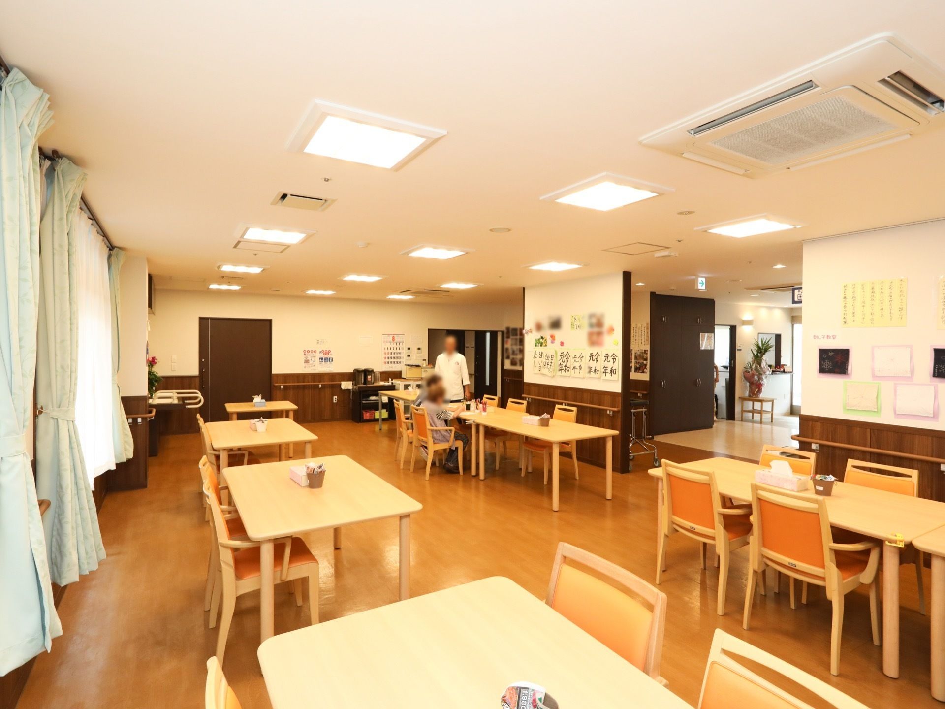 食堂の椅子に入居者様が２名座っている。その横に男性が１名立っている。天井には埋め込み式の照明器具が多く設置されている。