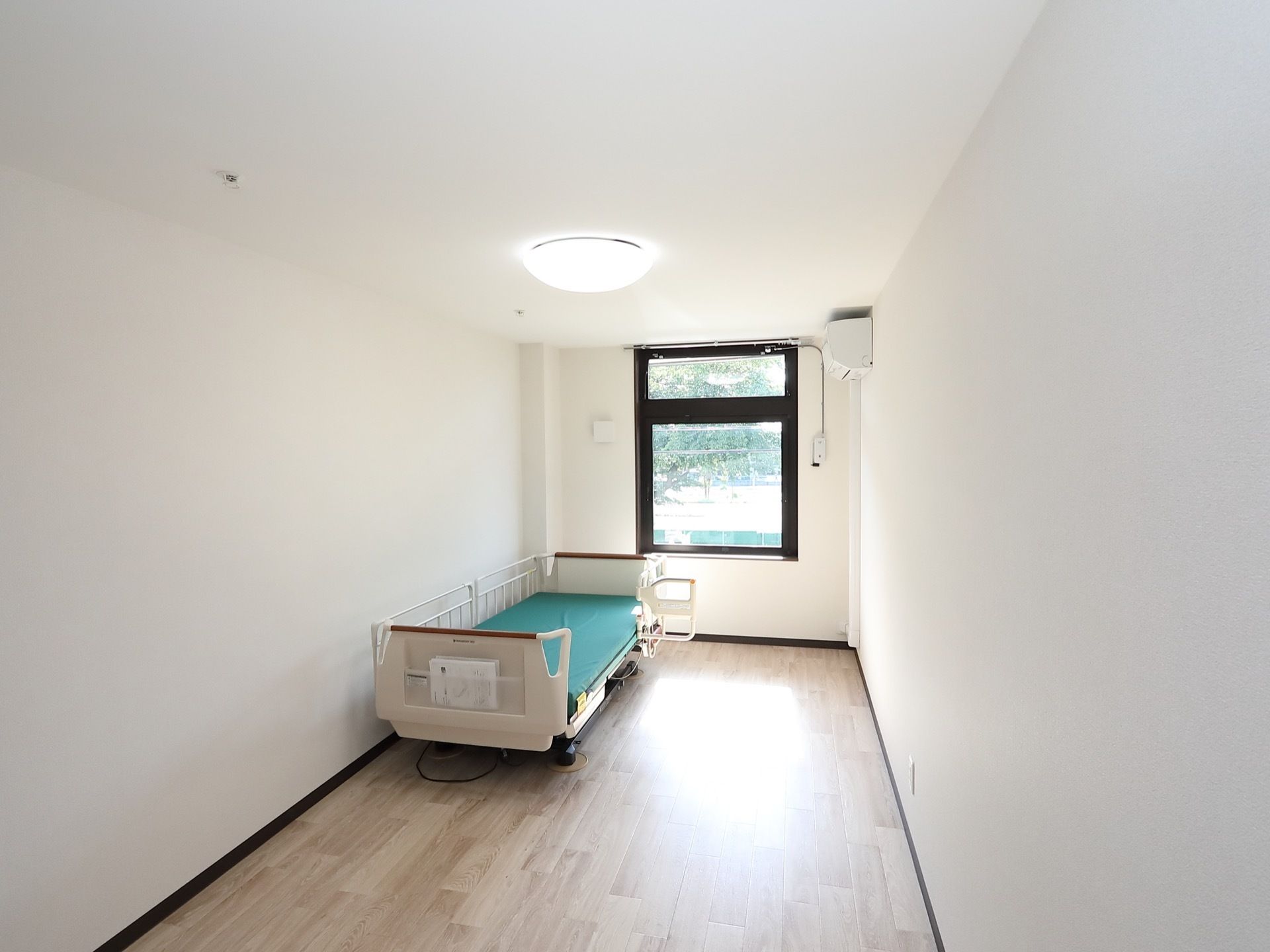 フローリングのシンプルな部屋に電動ベッドが１台設置されている。窓からは外の景色が見え、陽射しも良く入っている。