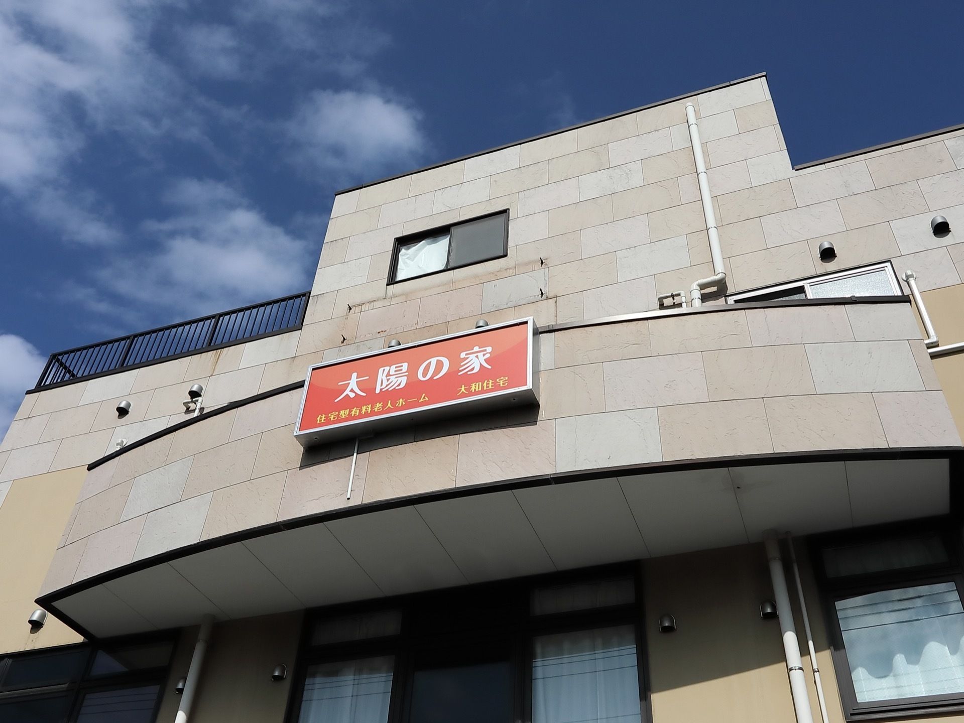 オフホワイトやグレーのタイルで彩られたマンションタイプの建物がある。正面には、ホームの看板が掲げられている。