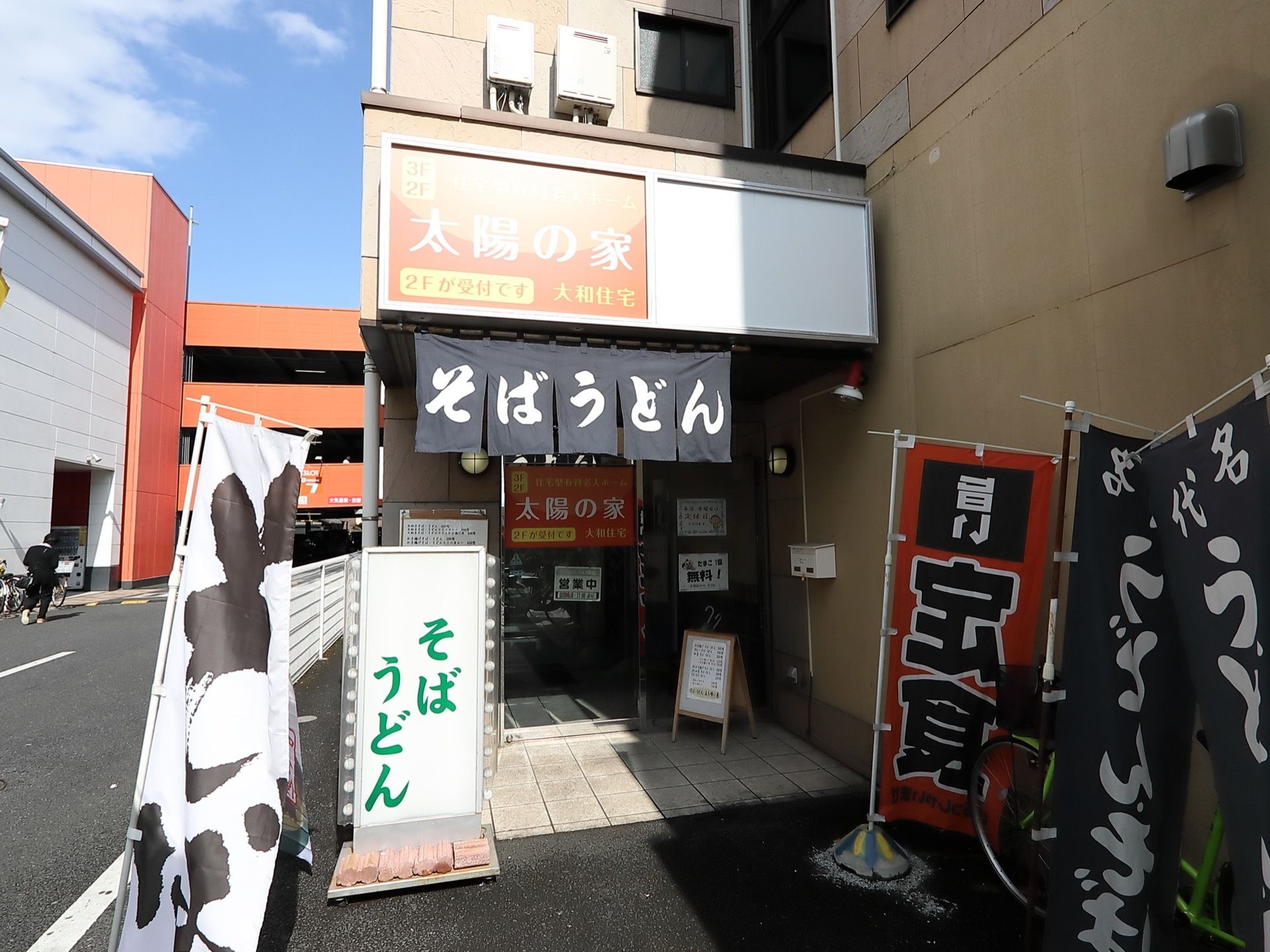 建物の１階部分は定食屋になっていて、のぼりや看板が出ている。軒や自動ドアにホームの案内が書かれている。