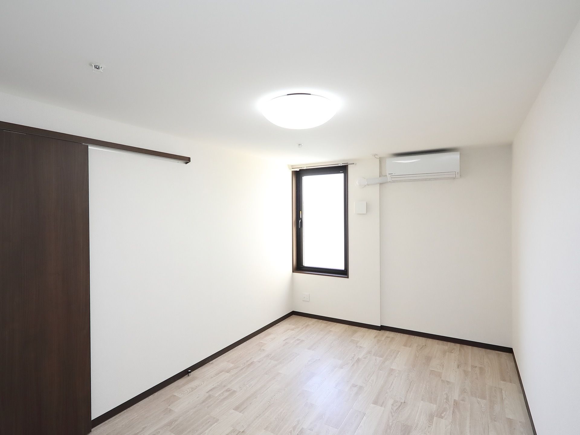 落ち着いた色のフローリングが敷かれた部屋がある。壁にはエアコンが設置されていて、腰の高さあたりに窓がついている。