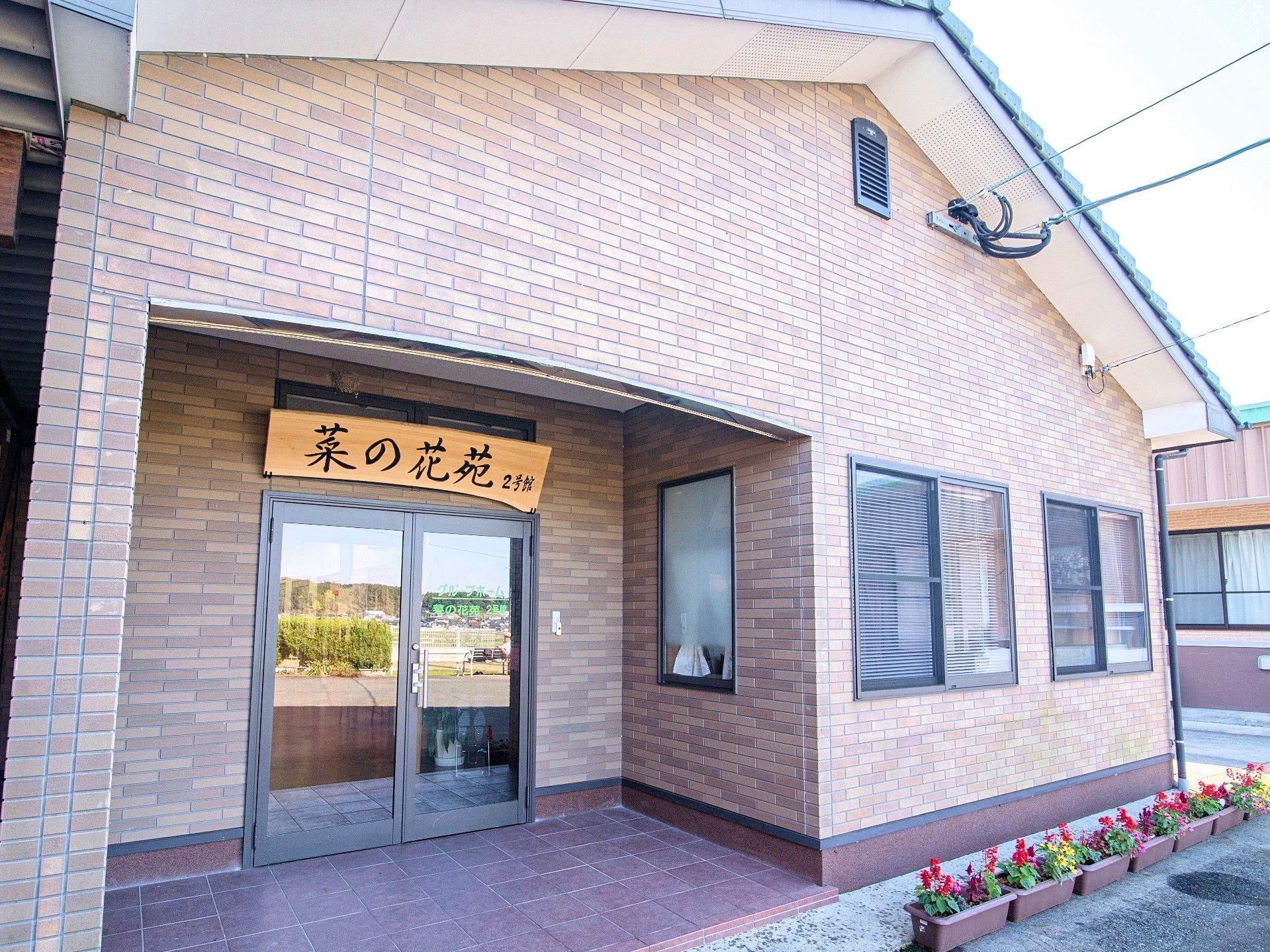 茶色い壁と三角形の屋根の建物。入り口には木製の板に施設名が書かれた看板がかかっている