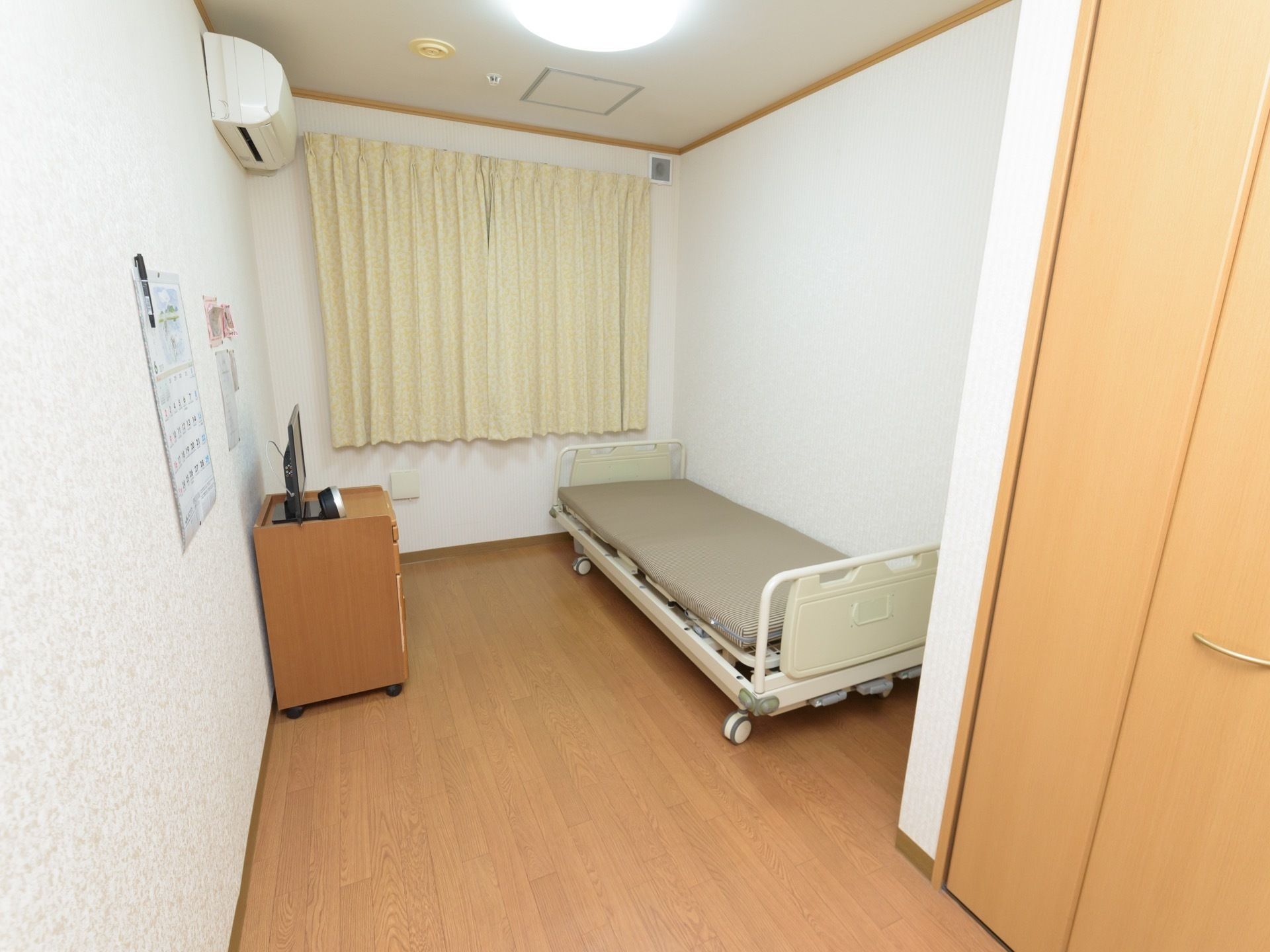 部屋の端に介護ベッドが置いてあり、腰高窓にクリーム色のカーテンが掛けられている。壁にエアコンが設置されている。