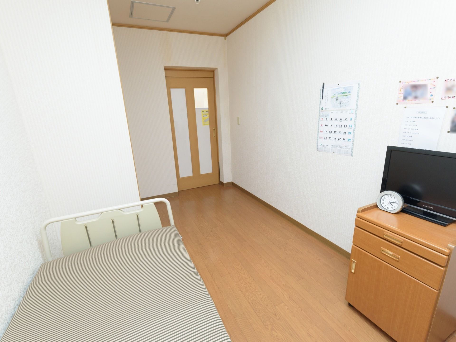 部屋の壁際に木目の台に乗せられた黒い薄型テレビが置いてある。壁にカレンダーや数枚の紙が貼られている。