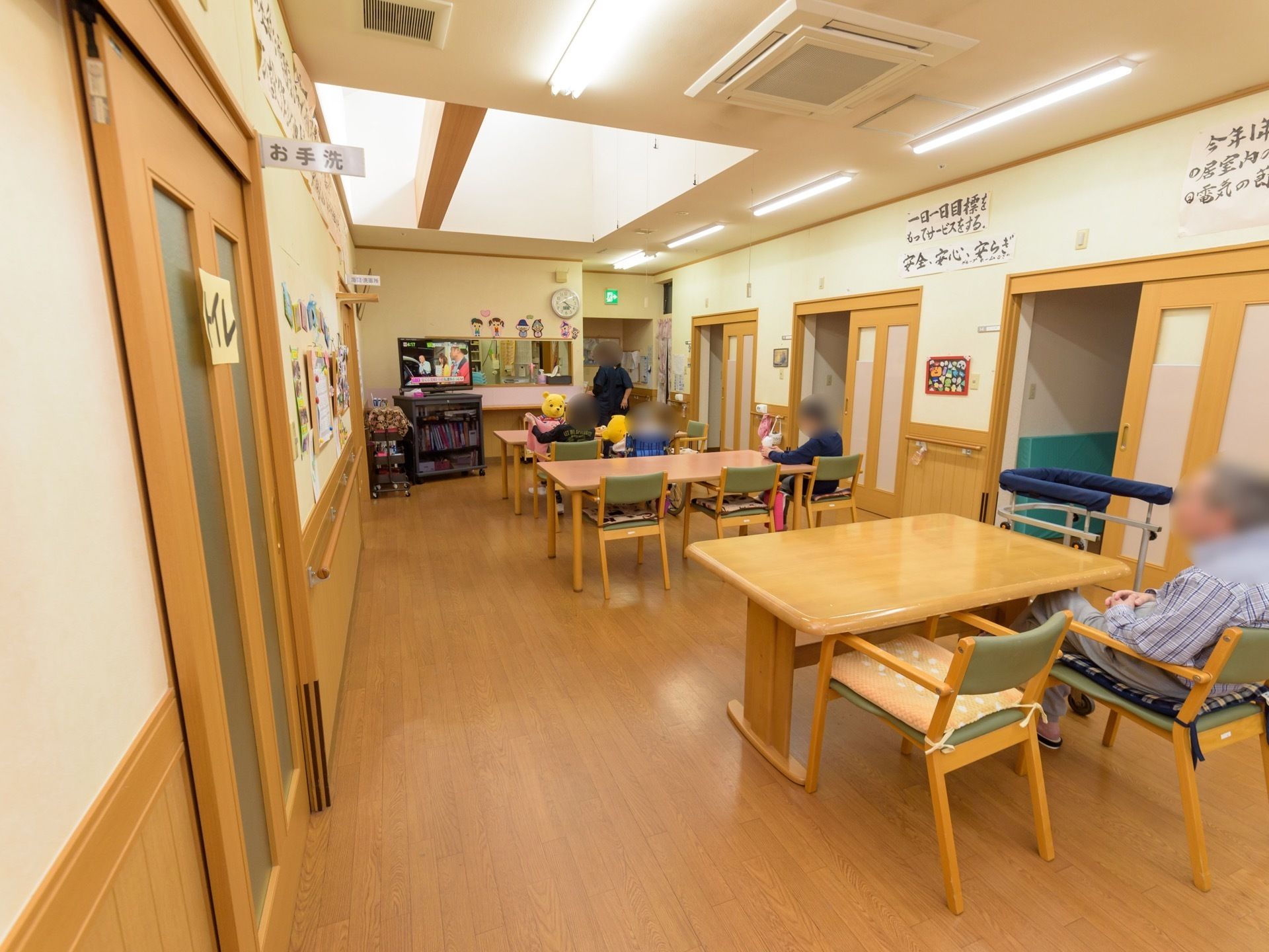 フローリングの床の細長い部屋にダイニングテーブルや椅子が置いてある。部屋の奥に設置されたテレビに人物が映っている。