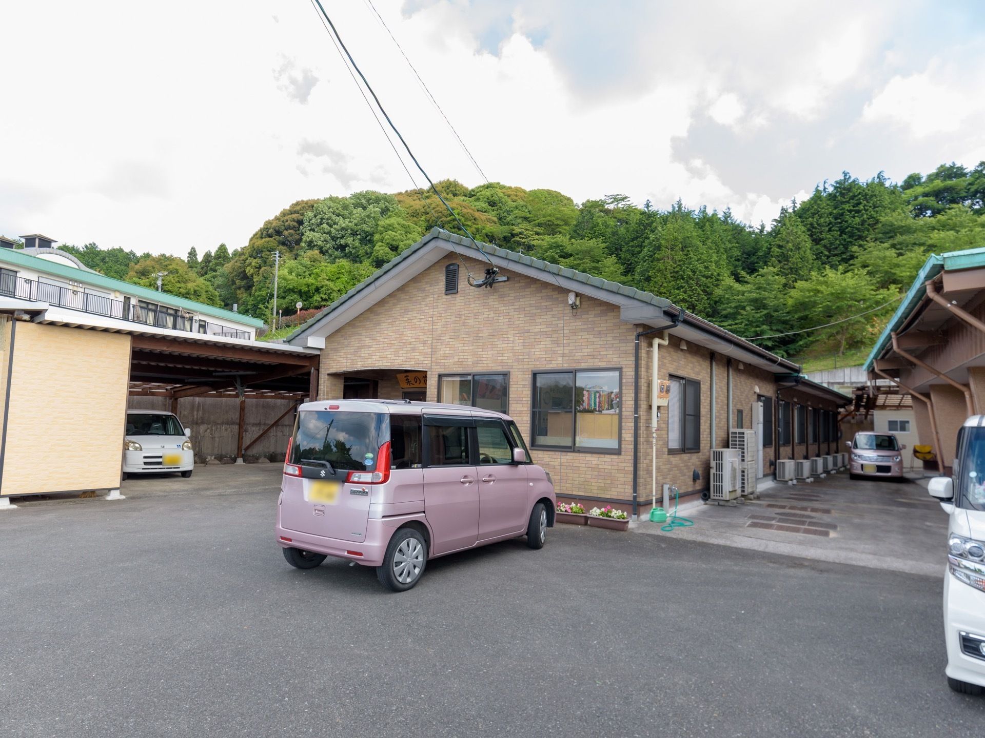 茶色い平屋建ての建物の向こう側に木々が生い茂っている。敷地内に数台の車が停められ、建物の脇にエアコンの室外機がたくさん置いてある。
