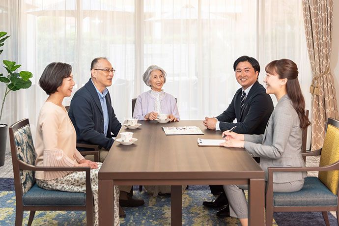 屋外のベンチに腰かける高齢のご夫婦に笑顔で話し掛ける２名の女性スタッフ