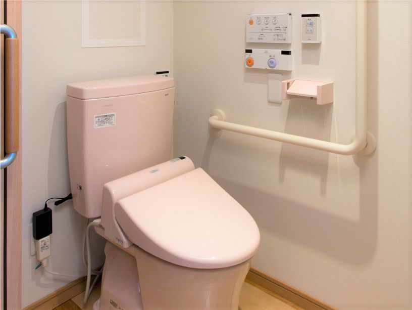 ピンクのトイレは温水便座が付いていて、壁にはエル字型の手すり屋操作パネル、トイレットペーパーホルダーが設置されている。