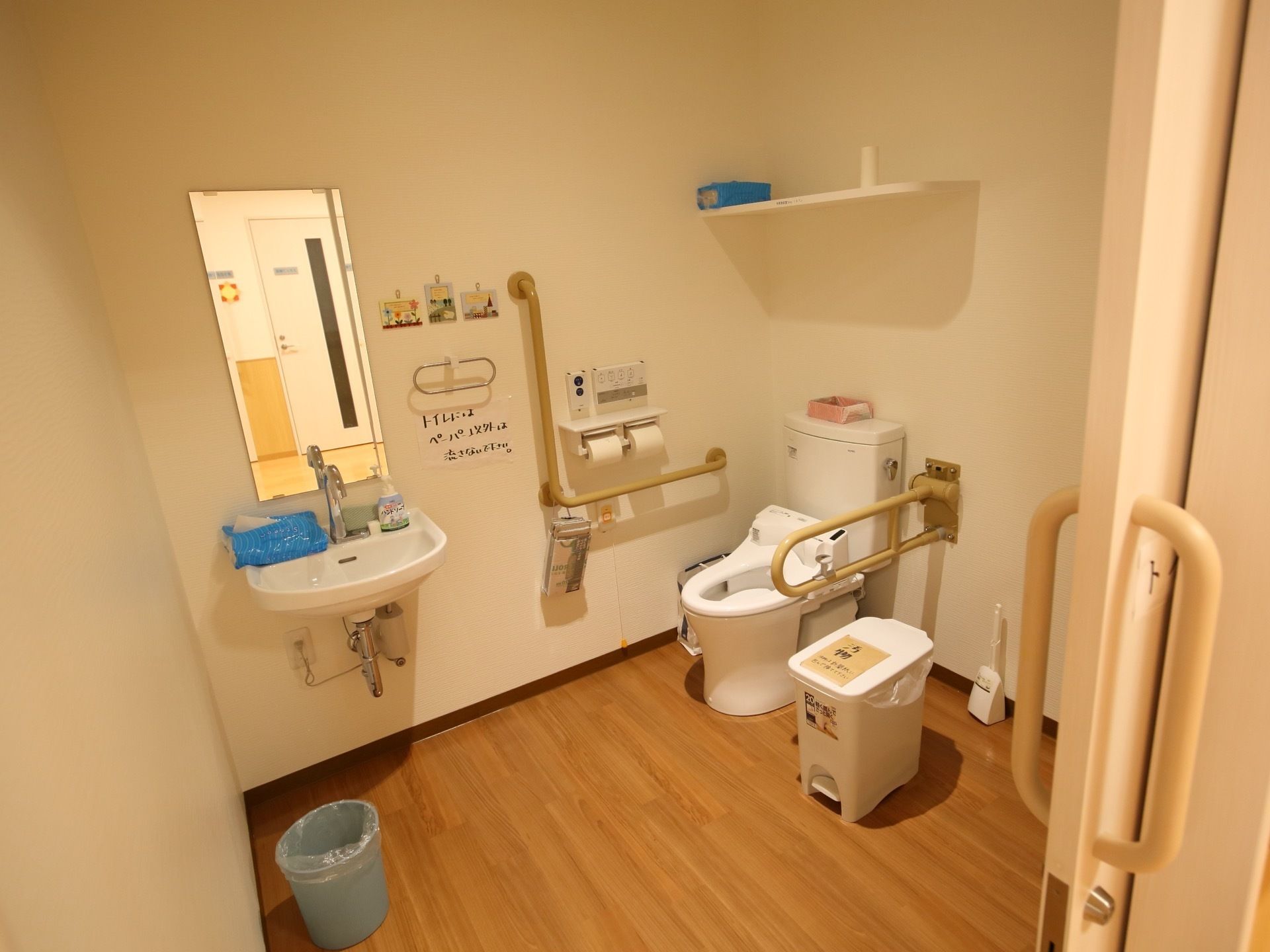 フローリングの広いトイレがある。便座の周りや壁には手すりが設置されていて、鏡や洗面設備も付いている。