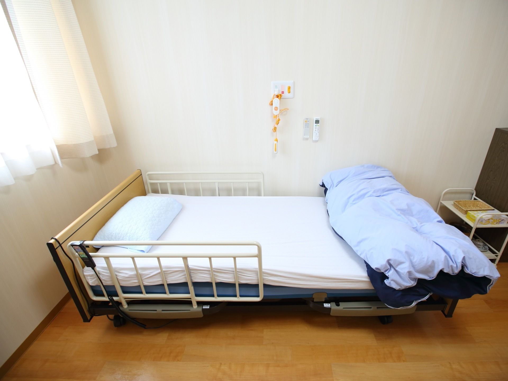 居室の窓際に電動ベッドが置かれている。壁には照明や空調のリモコンのほか、緊急時の通報装置が設置されている。