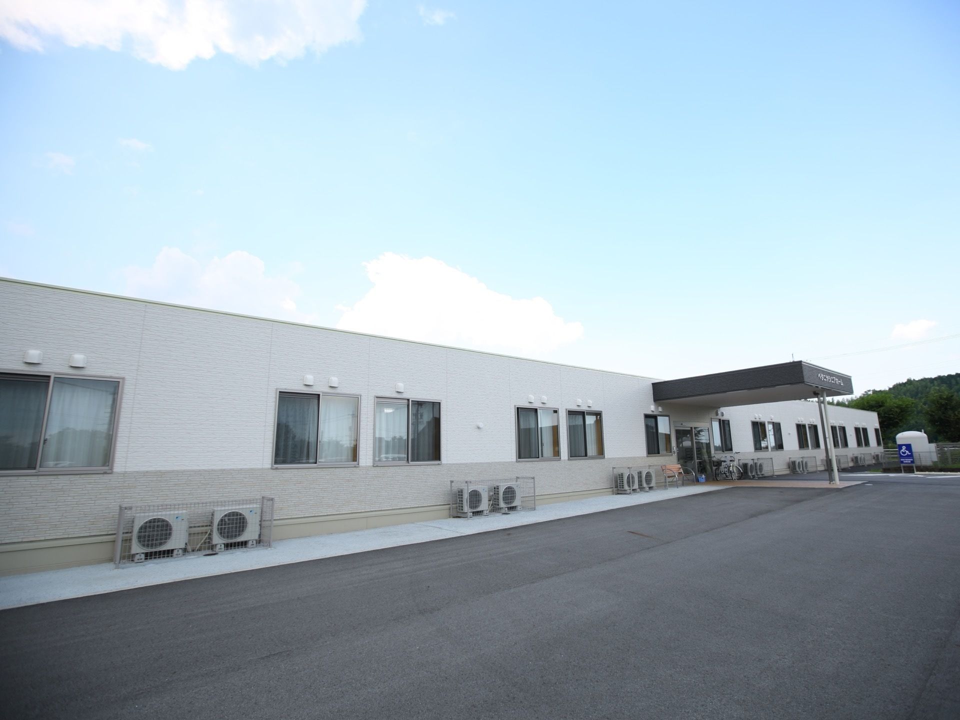 アスファルトで舗装された広い敷地に、長い平屋の建物が建っている。玄関部分には軒が付いていて、近くに車椅子用の駐車スペースがある。