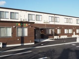 白とブラウンの2階建ての建物の前には駐車場があり、2階部分の外壁にはホーム名が書かれている。