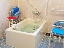 白い一人用の浴槽が設置され、可動式の手すりが浴槽に、固定式の手すりが壁に付き、シャワーチェアも置かれている。