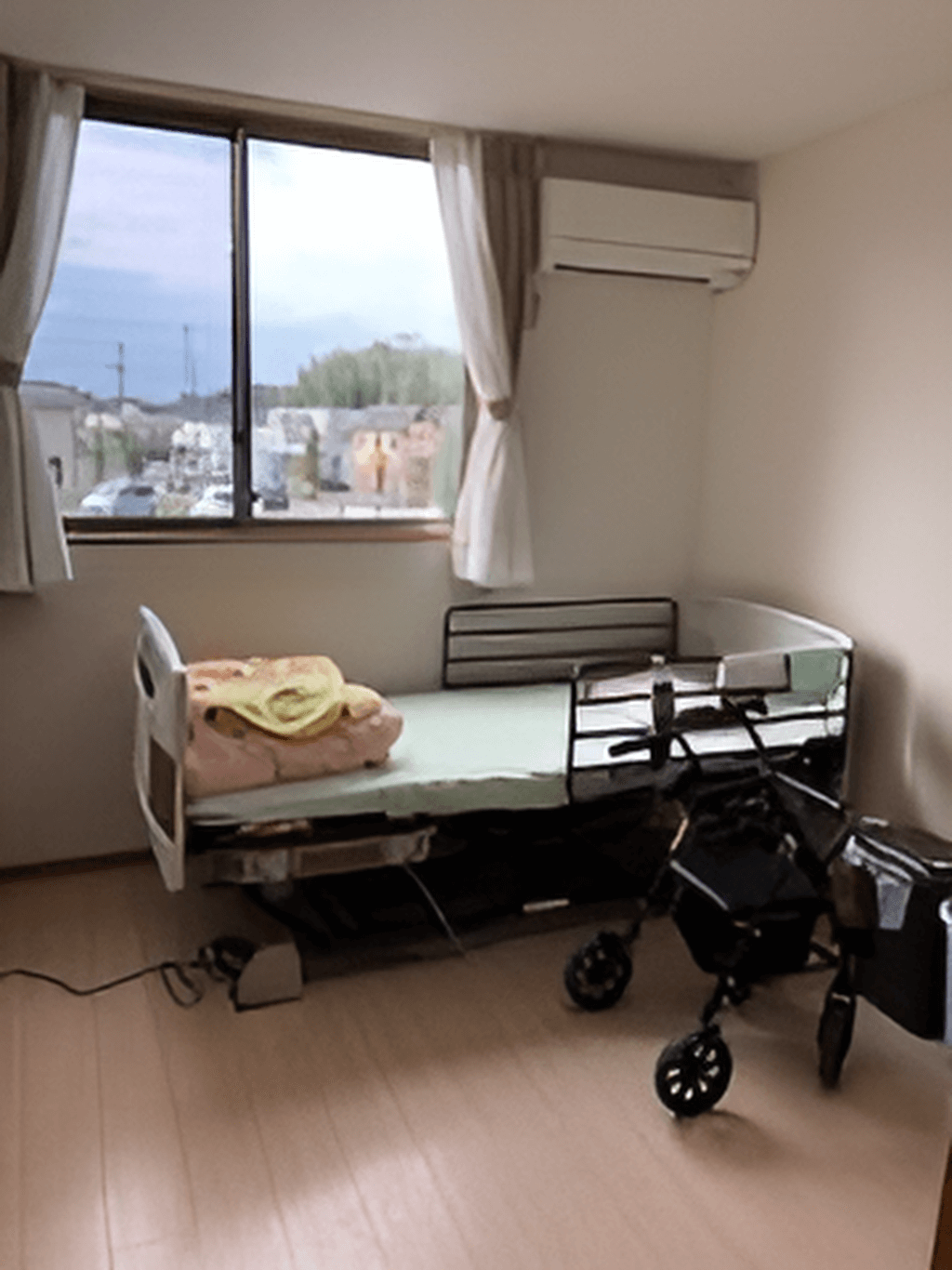 カーテンが付いた大きな窓があるフローリングの部屋にはベッドや車いすがあり、エアコンもついている。