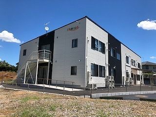 ライトグレーとダークグレーの2階建ての建物の前には駐車場があり、自然に囲まれている。