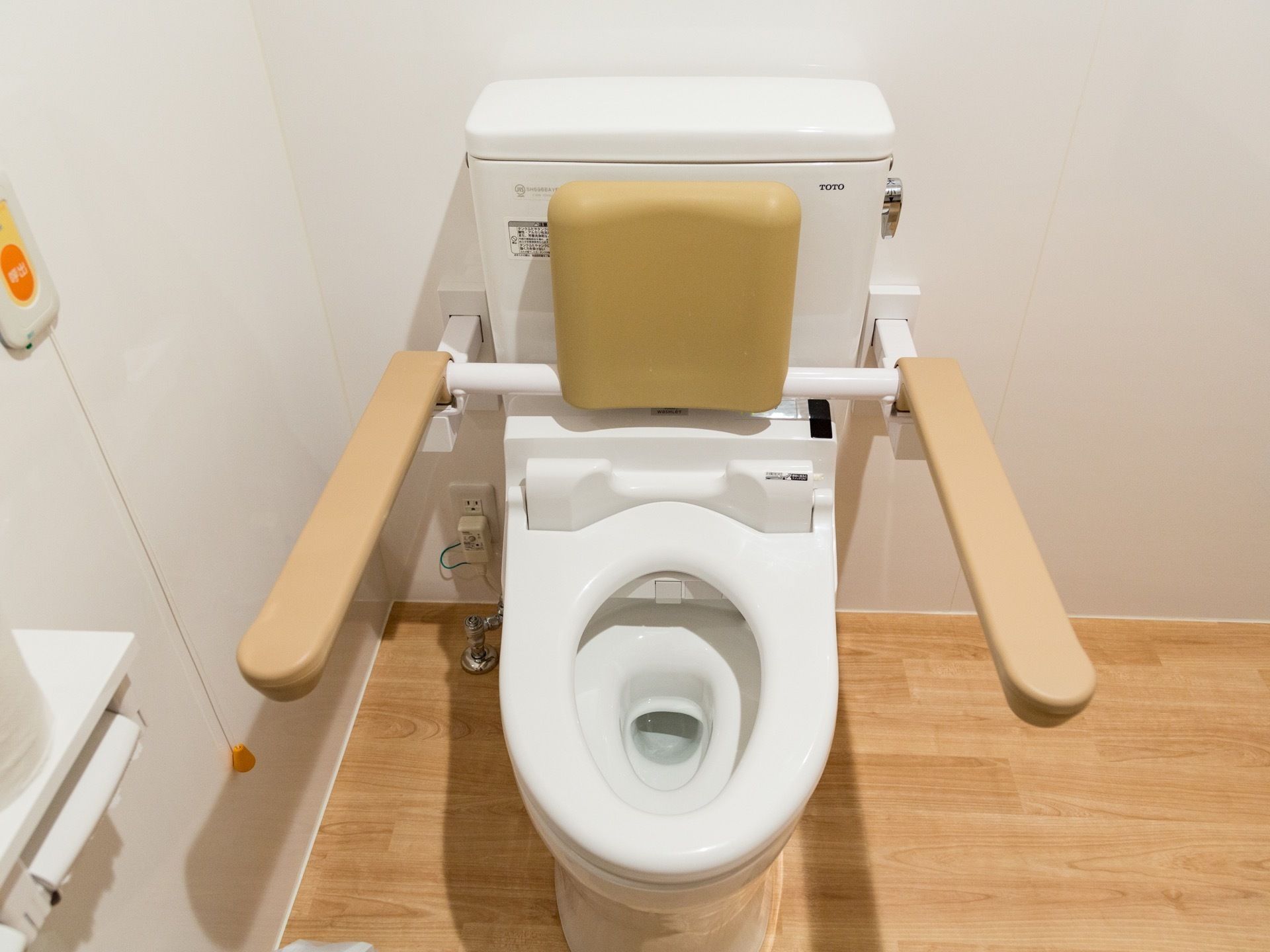 トイレの便座には、背もたれと肘掛状のパーツがセットになった器具を取り付けている。壁にオレンジ色のボタンの呼び出しベルがある。