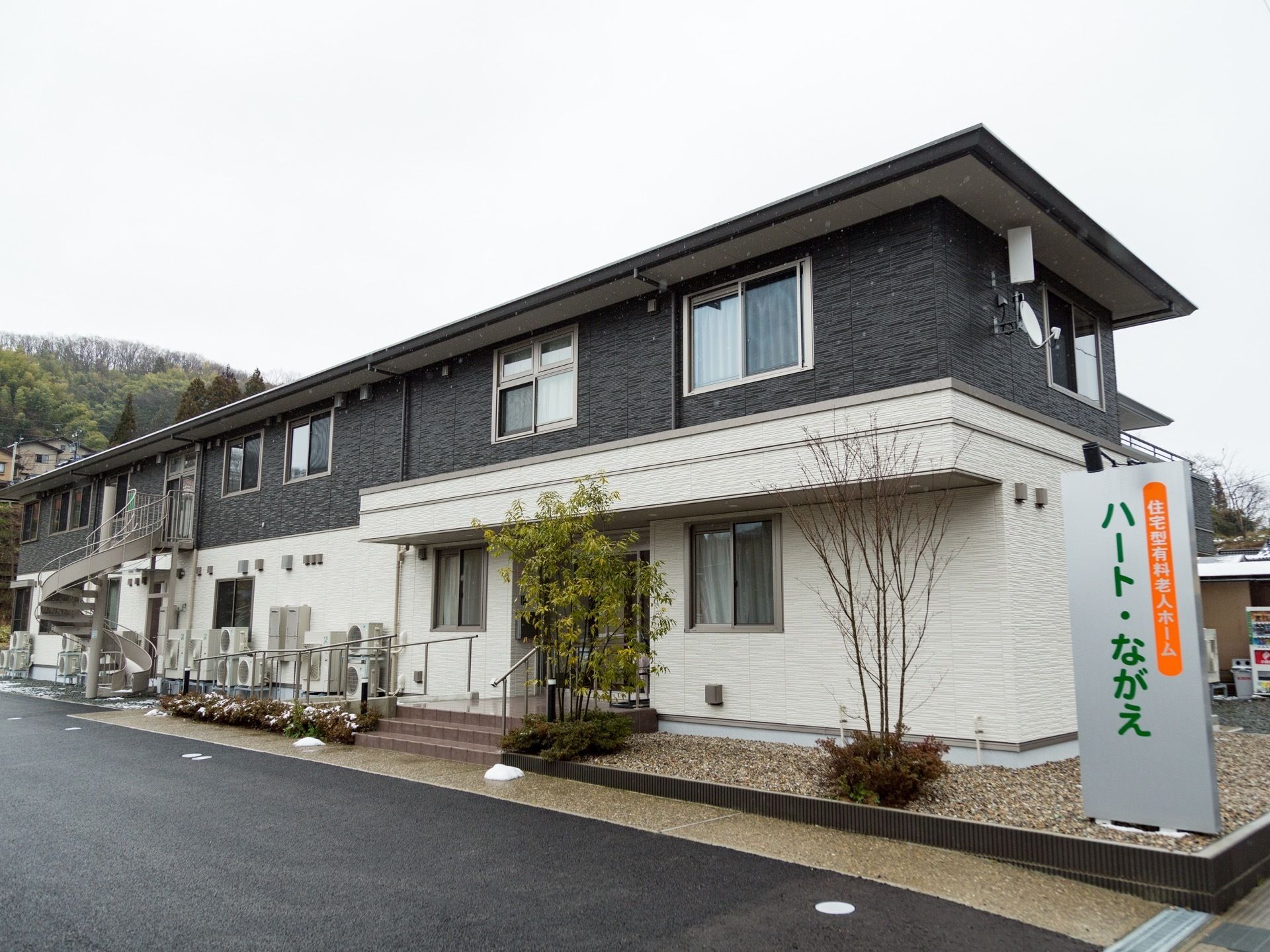 正面玄関とは別の場所に、２階から螺旋階段が設置することで、避難経路を確保している。道路側に銀色の看板が出ている。