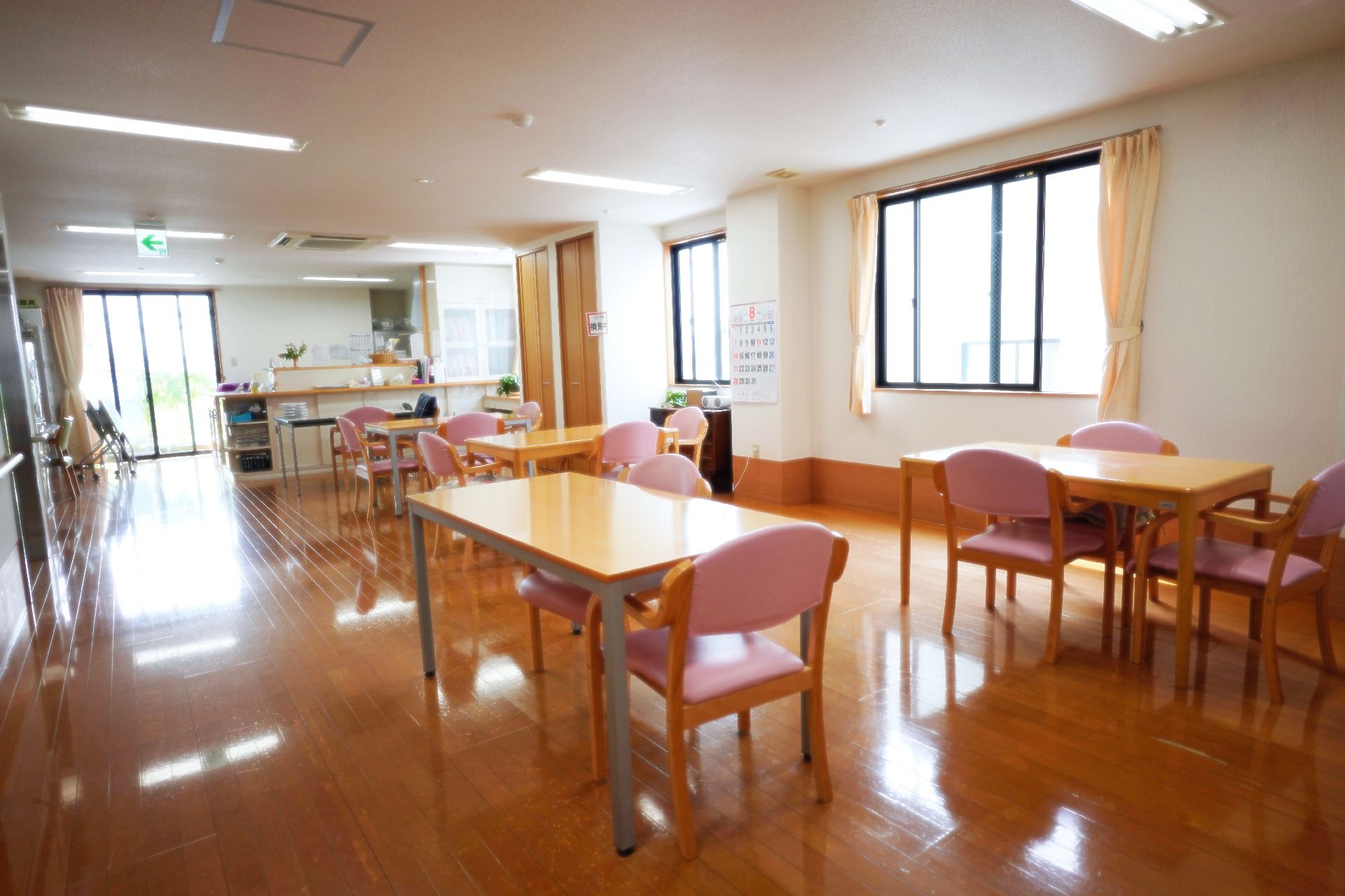 木製の机とピンクのクッションが付いた椅子が並んでいる白い壁の広間