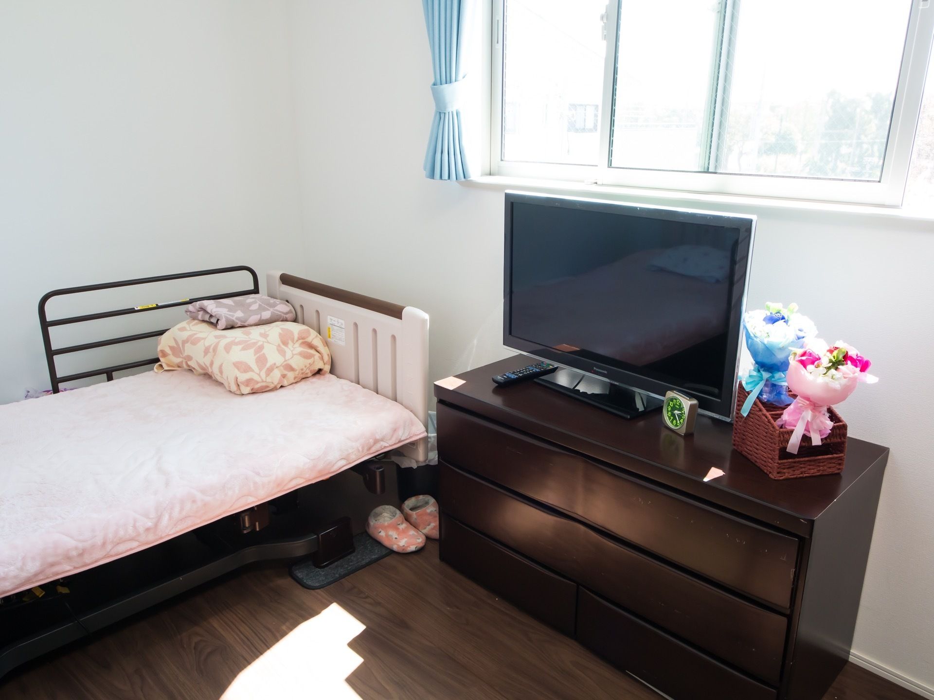 居室に、窓から日が差し込んでいる。窓際に置かれた衣類タンスの上にテレビが載っていて、その前には置き時計がある。
