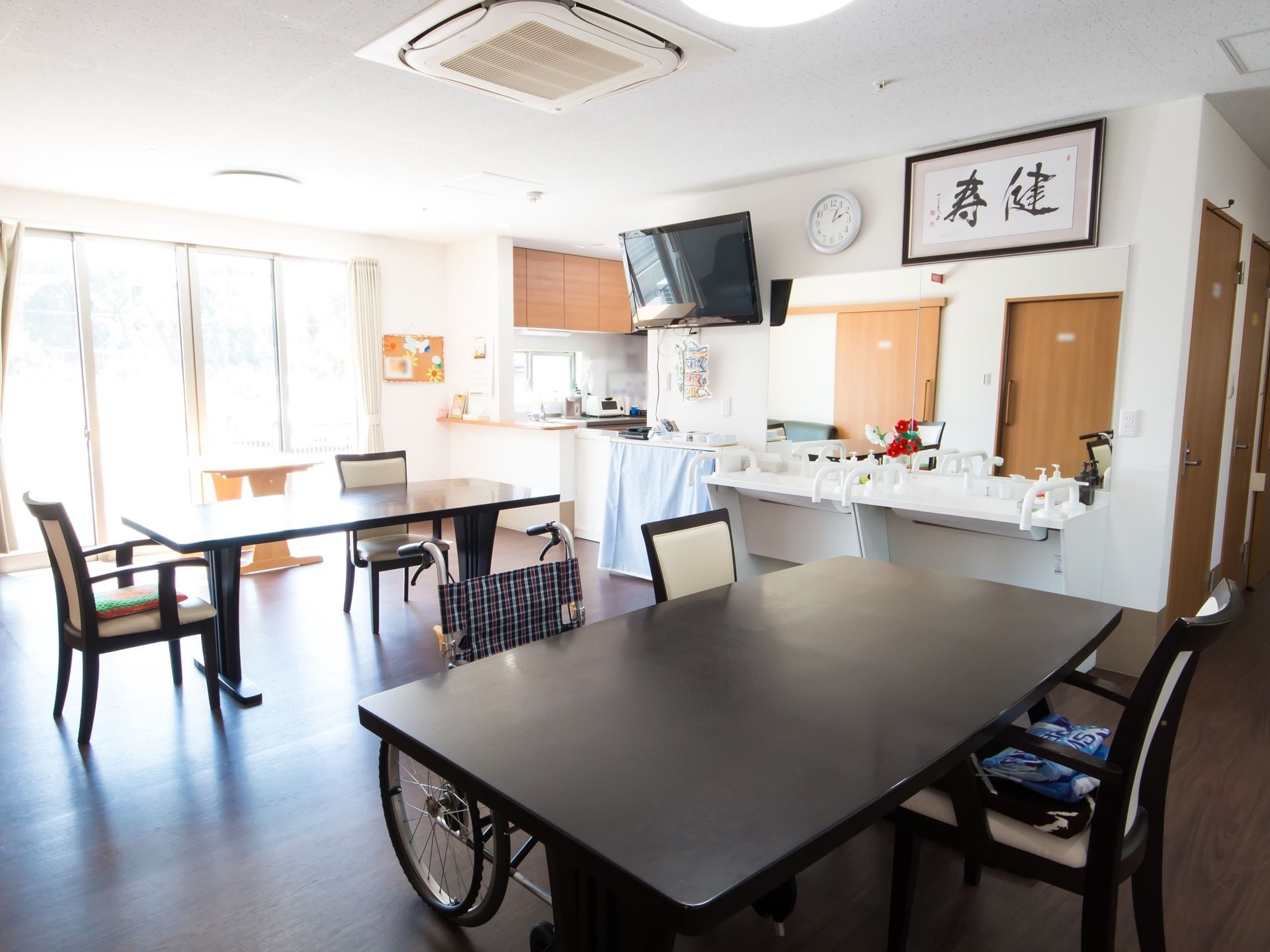 食堂にはダイニングテーブルと椅子が用意されていて、空いている席には車椅子が置かれている。壁には洗面台と鏡が設置され、テレビも掛けられている。