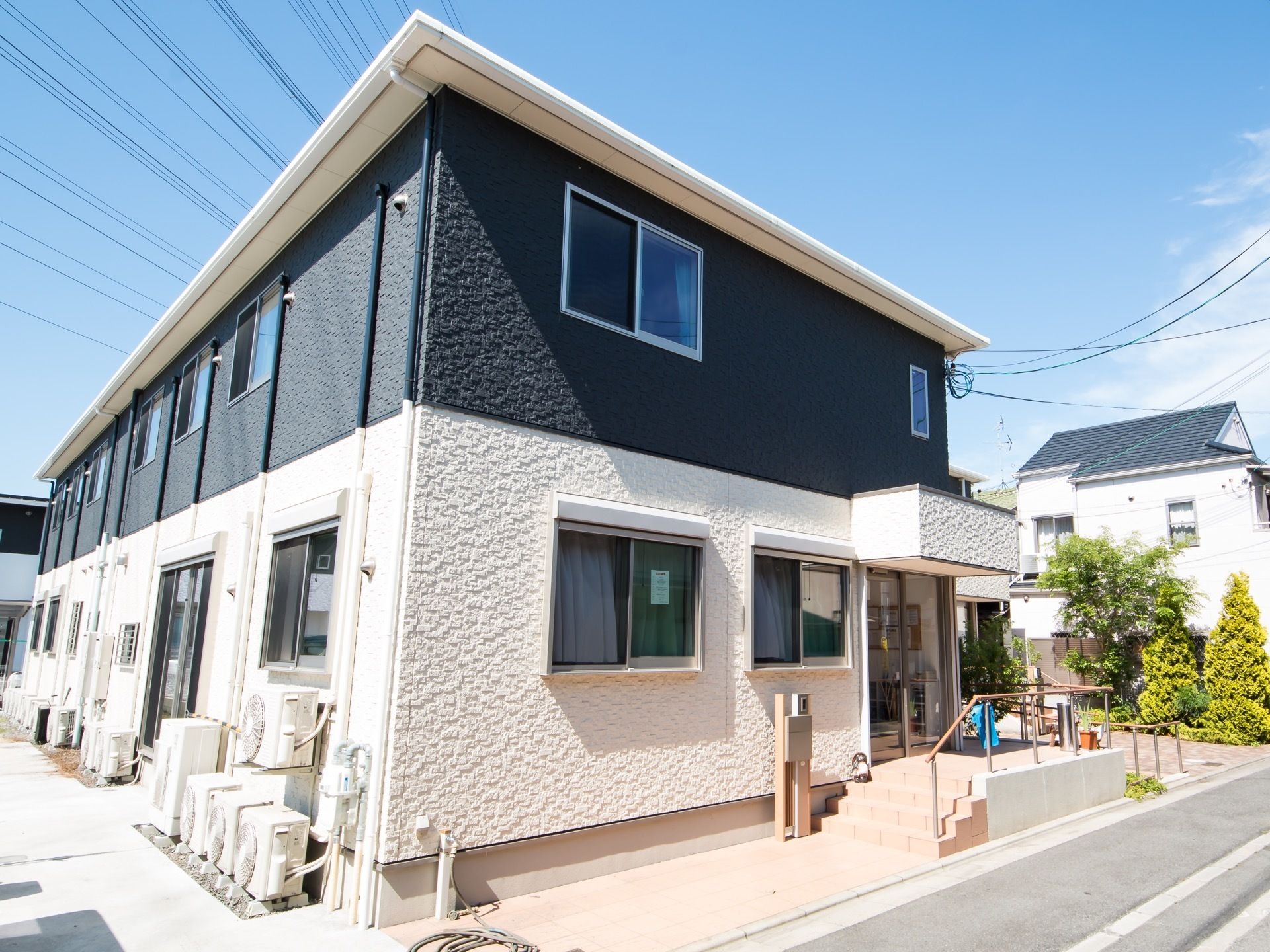 建物は2階建てで、1階は白、2階は濃いグレーの外壁になっている。壁際にはエアコンの室外機が数台設置されている。