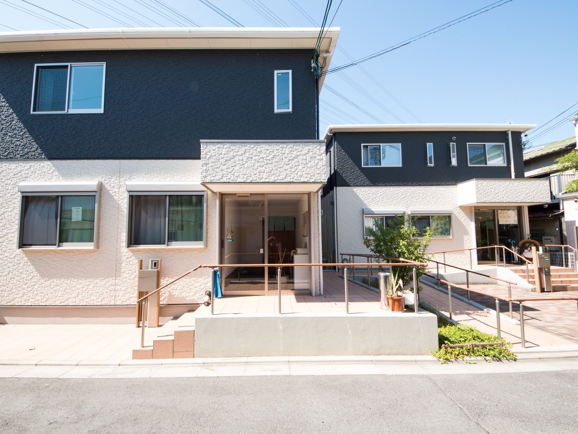 1階部分が白色で、2階部分が紺色に塗装された1戸建てが2棟並んでいる。それぞれ玄関までは階段とスロープがある。