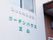 白を貴重とした壁に、緑色とグレーの中ぐらいの大きさの文字で施設の名前が示されている。その側には玄関がある。